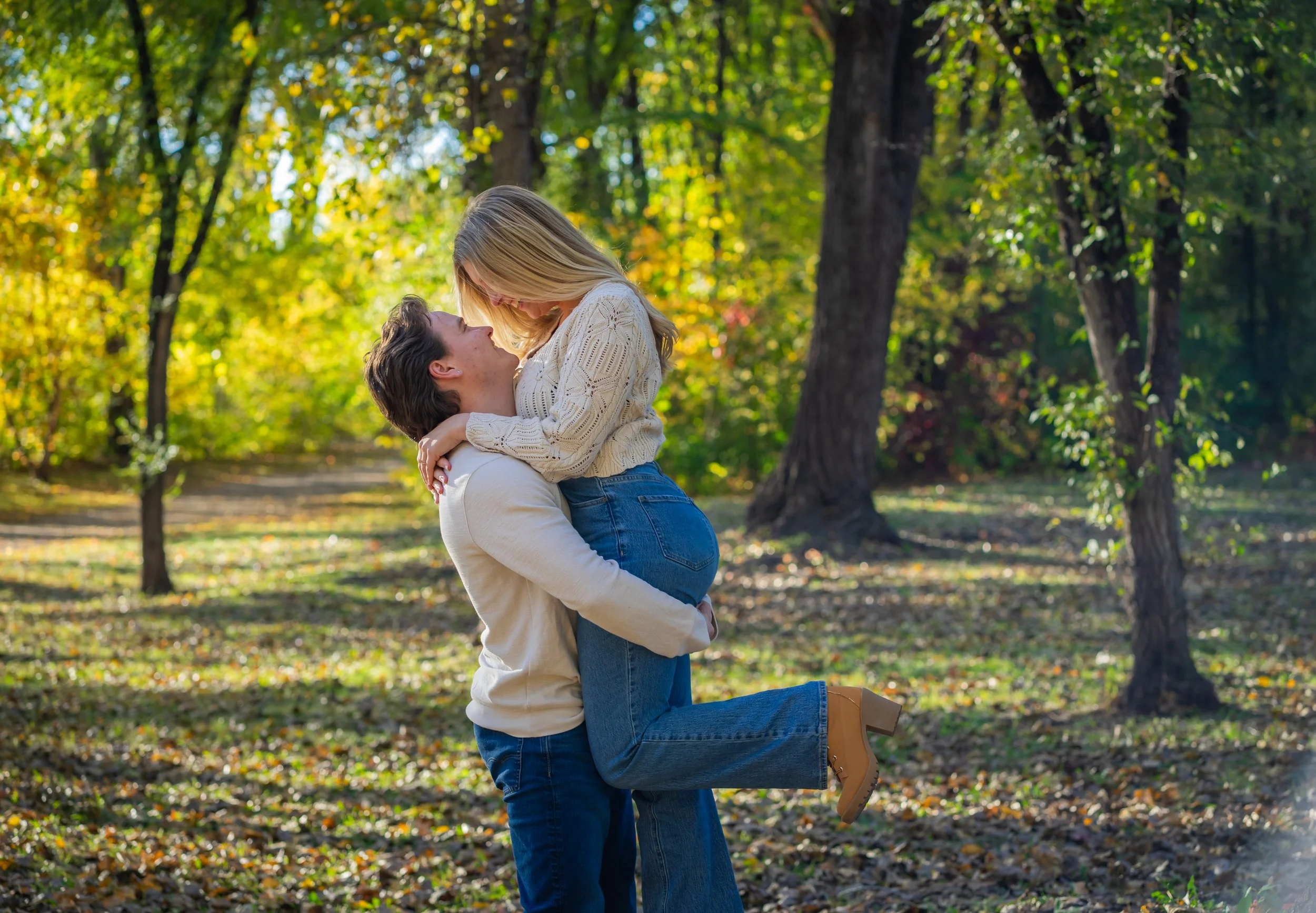 A man kneeling and holding a woman in the air, both smiling in a park with trees and autumn leaves.