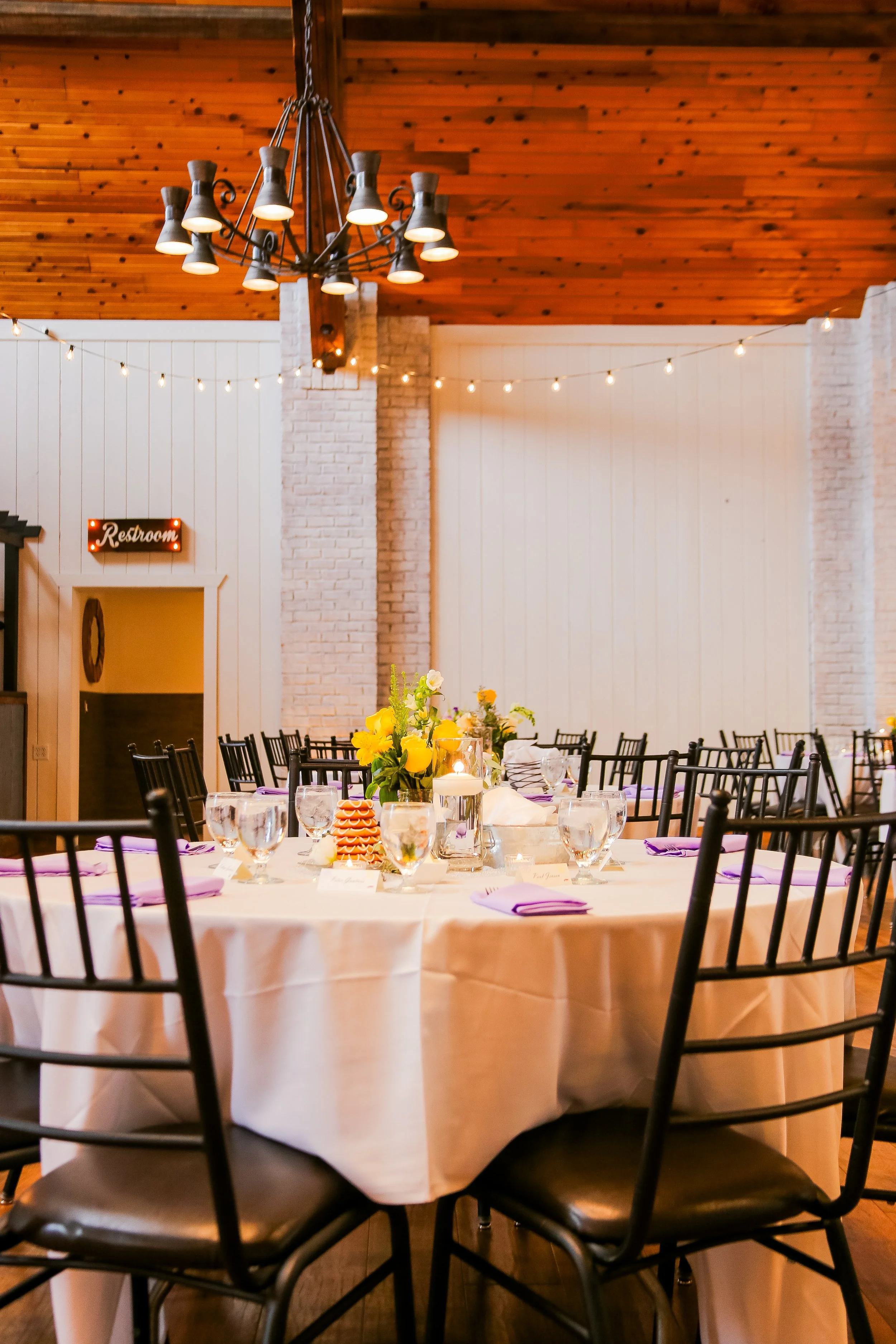 A decorated banquet table set for a meal, with flowers, candles, and purple napkins, in a warmly lit event space with string lights and a wooden ceiling.