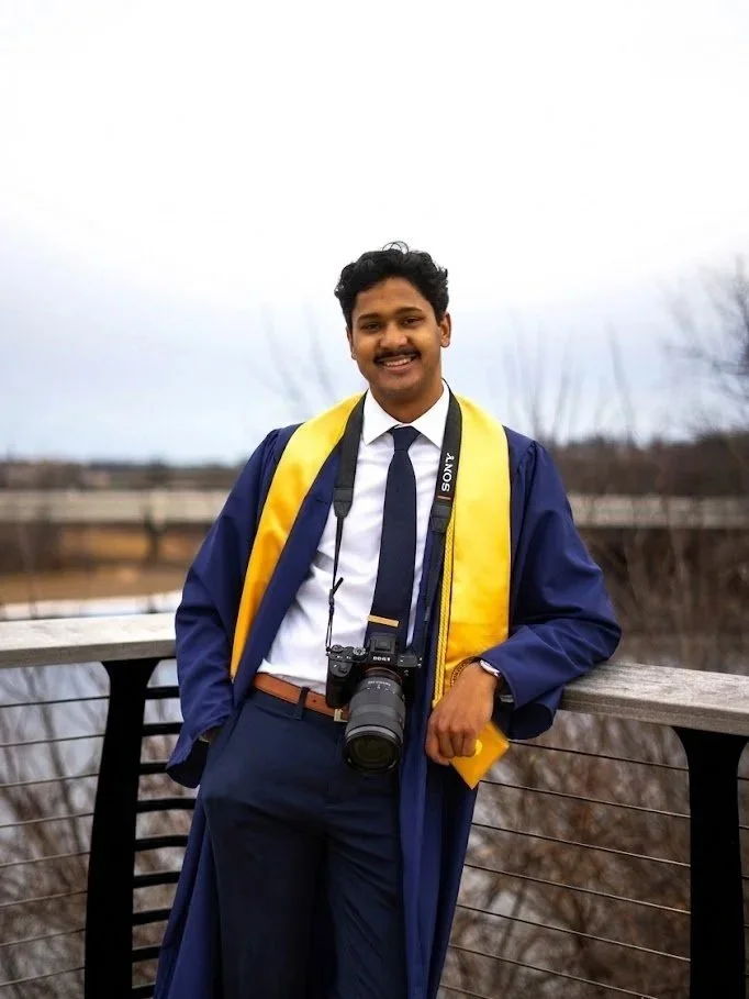 A young man in a blue graduation gown and yellow sash, holding a camera around his neck, standing outdoors on a bridge with a railing against a cloudy sky background.