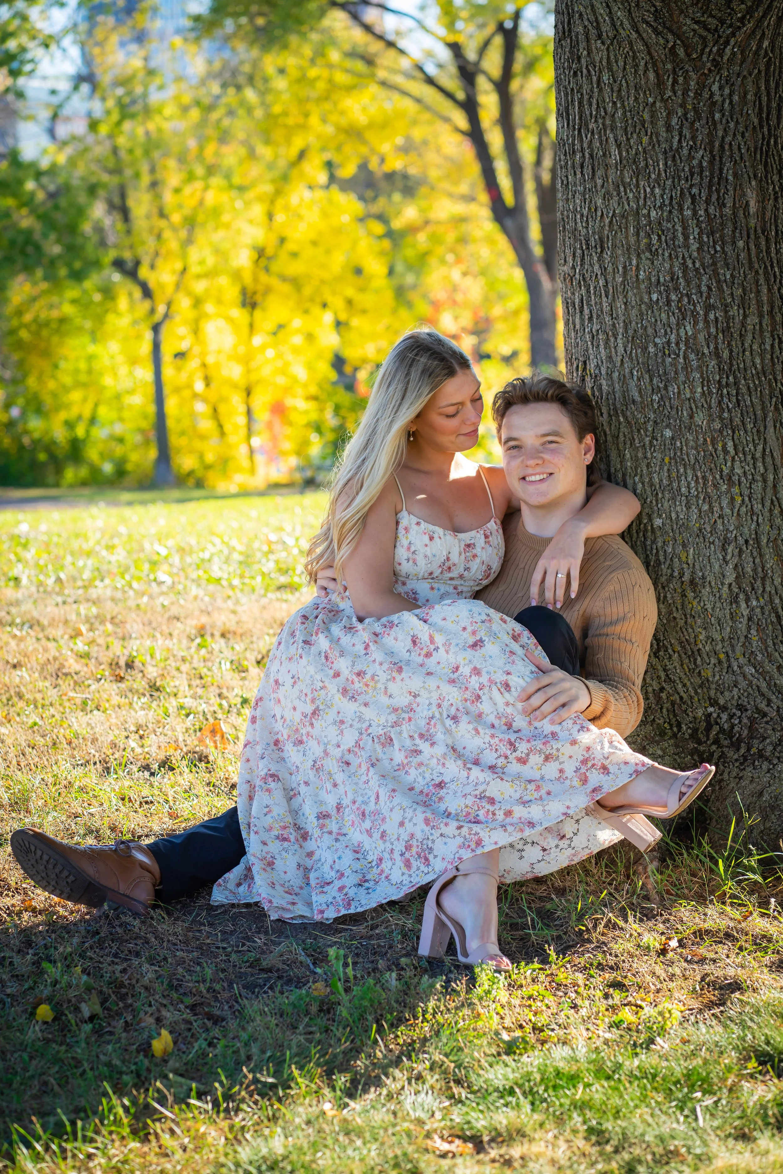 A young woman and man sitting on the grass and leaning against a large tree in a park during fall. The woman has long blonde hair and is wearing a floral dress, while the man has short brown hair and is dressed in a brown sweater and dark pants. They are smiling and close together, with the woman resting her arm on the man's shoulder.
