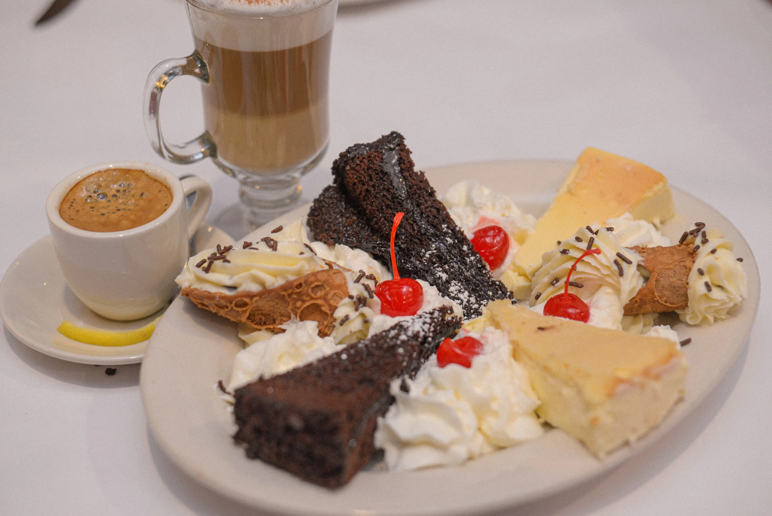 A plate of assorted desserts including cannolis, chocolate cake, and cheesecake, with a cup of espresso and cappuchino.