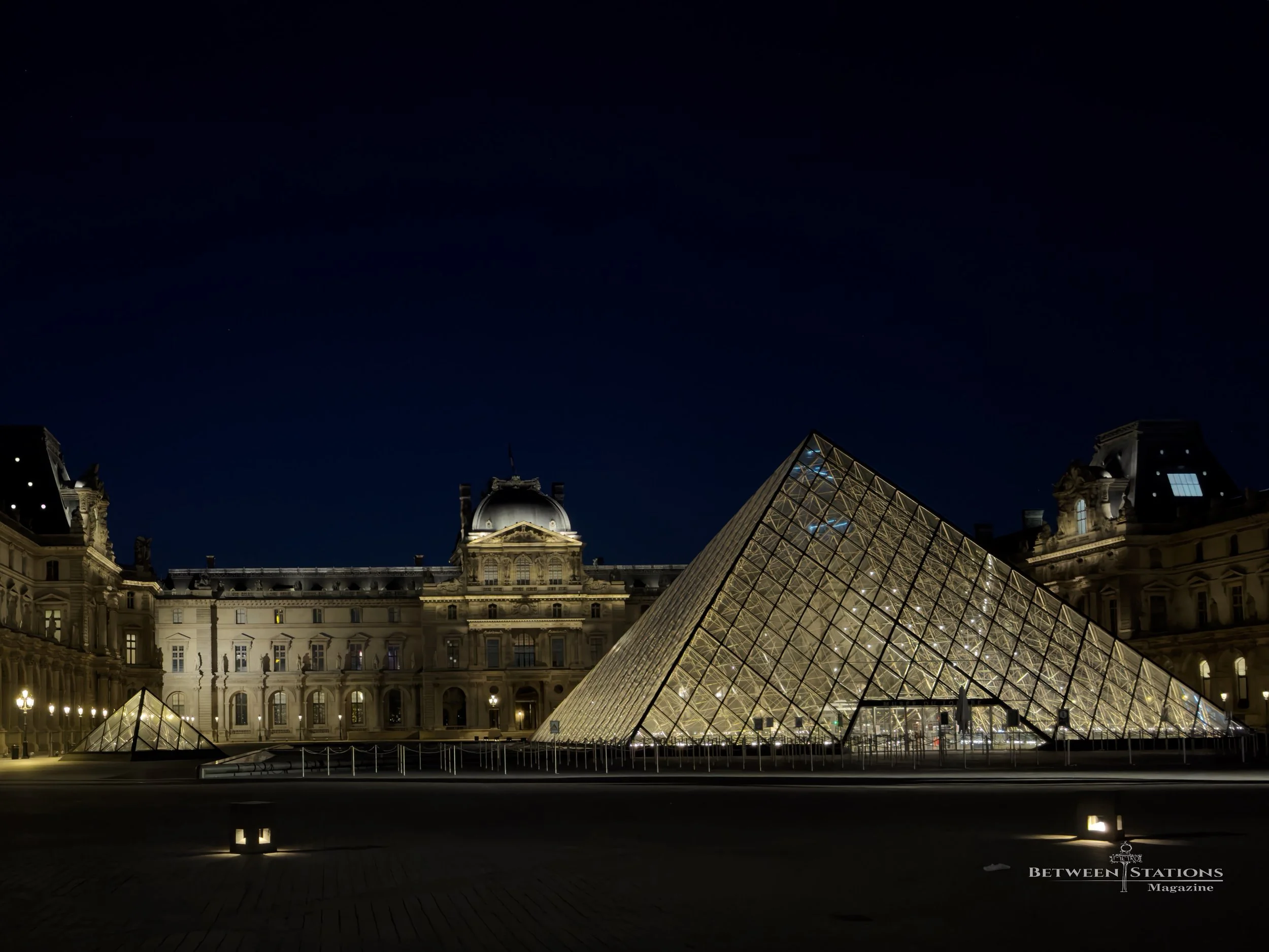 The Louvre Pyramid glows with warm light against a nearly black-blue night sky. Behind it, the museum's ornate facades are softly lit, with a domed pavilion at the center and mansard roofs on either side. A smaller pyramid sits at left, and rows of l