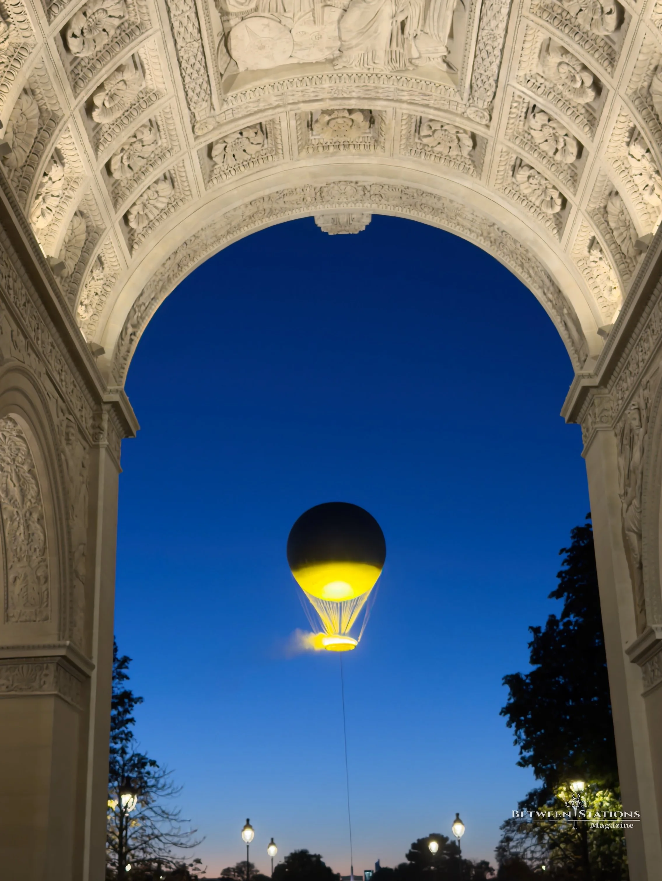 Looking up through an arch of richly carved stone, with a coffered ceiling of floral reliefs, the tethered Olympic cauldron hovers in the twilight sky. The balloon is dark at the top and lit bright yellow below, with its ring of light and a wisp of v