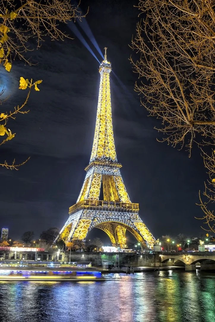 The tower lit gold against the night sky, its searchlight sweeping overhead while a passing boat streaks light across the water below.
