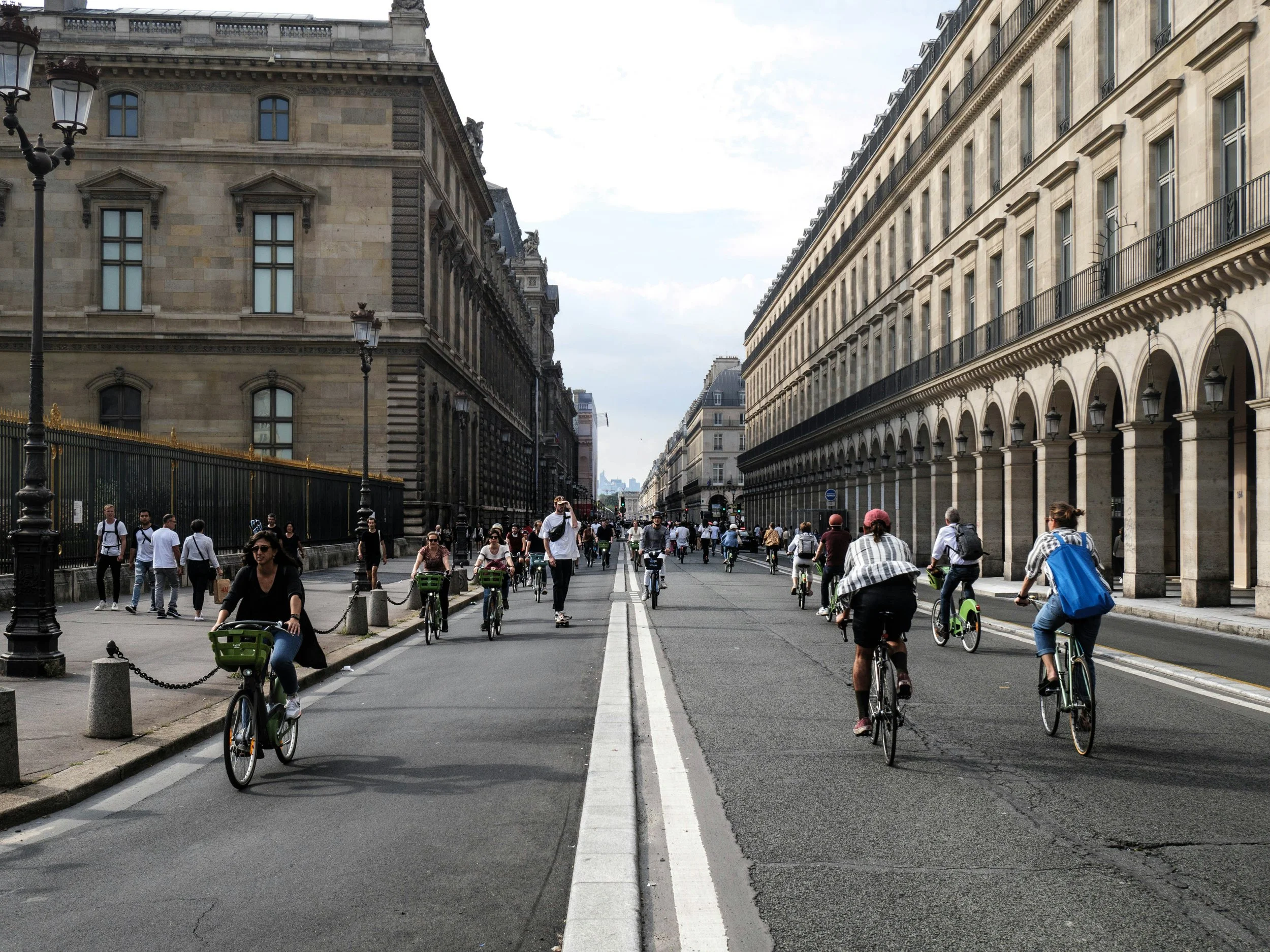 Large group of cyclists riding down a wide Paris boulevard alongside the Louvre's arcaded facade