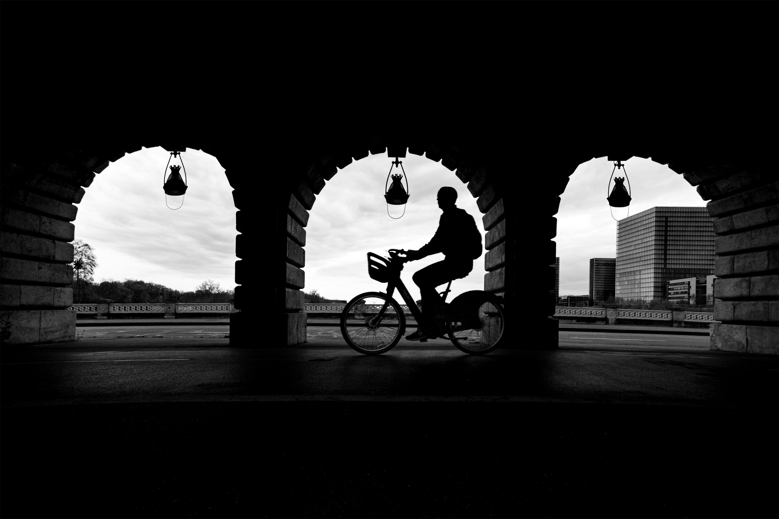 Silhouette of a cyclist riding beneath a stone archway with hanging lanterns, modern buildings visible beyond