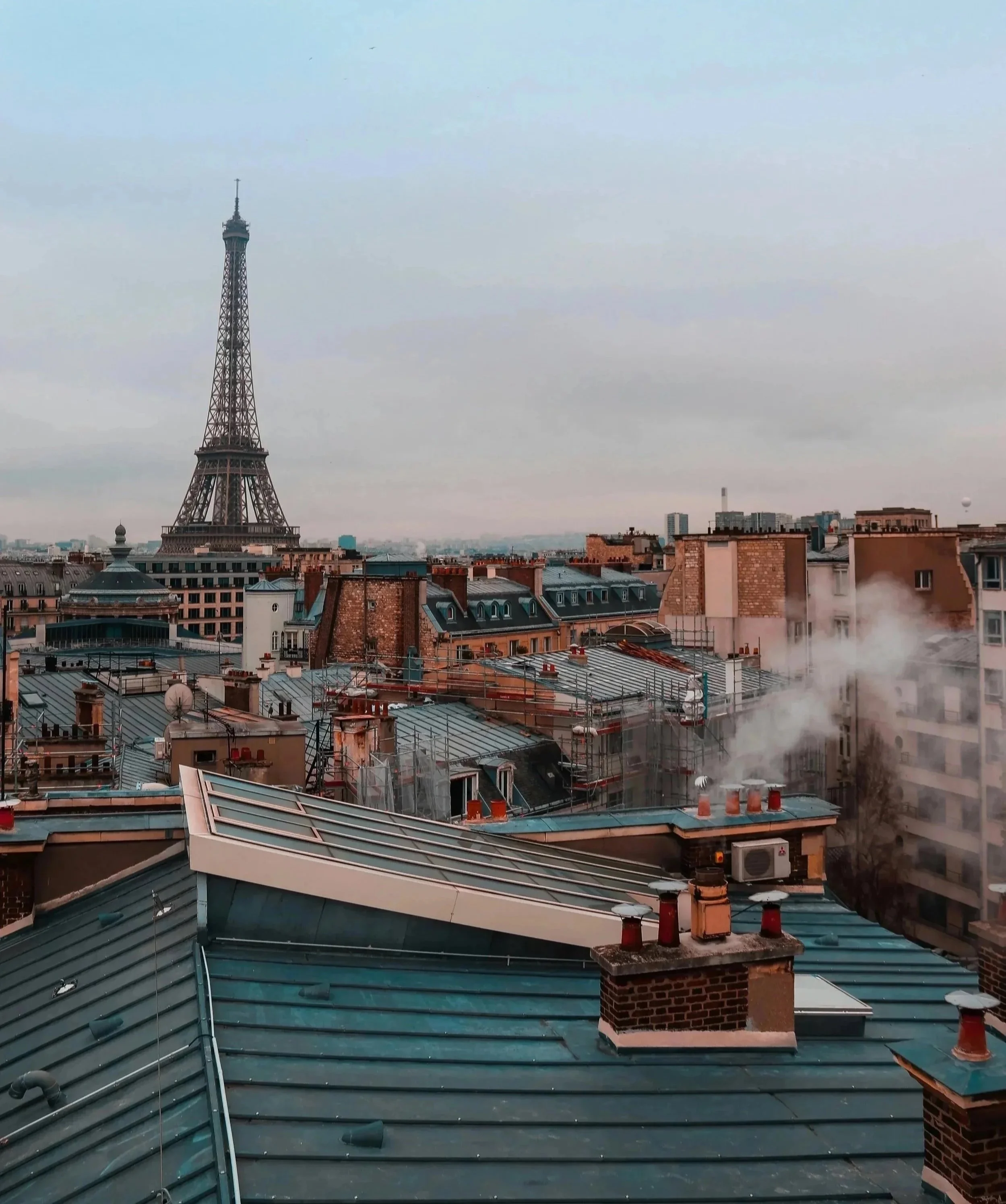 View of Paris skyline with the Eiffel Tower in the distance, seen from rooftops with chimneys and HVAC vents, and some smoke or steam rising from the rooftop.