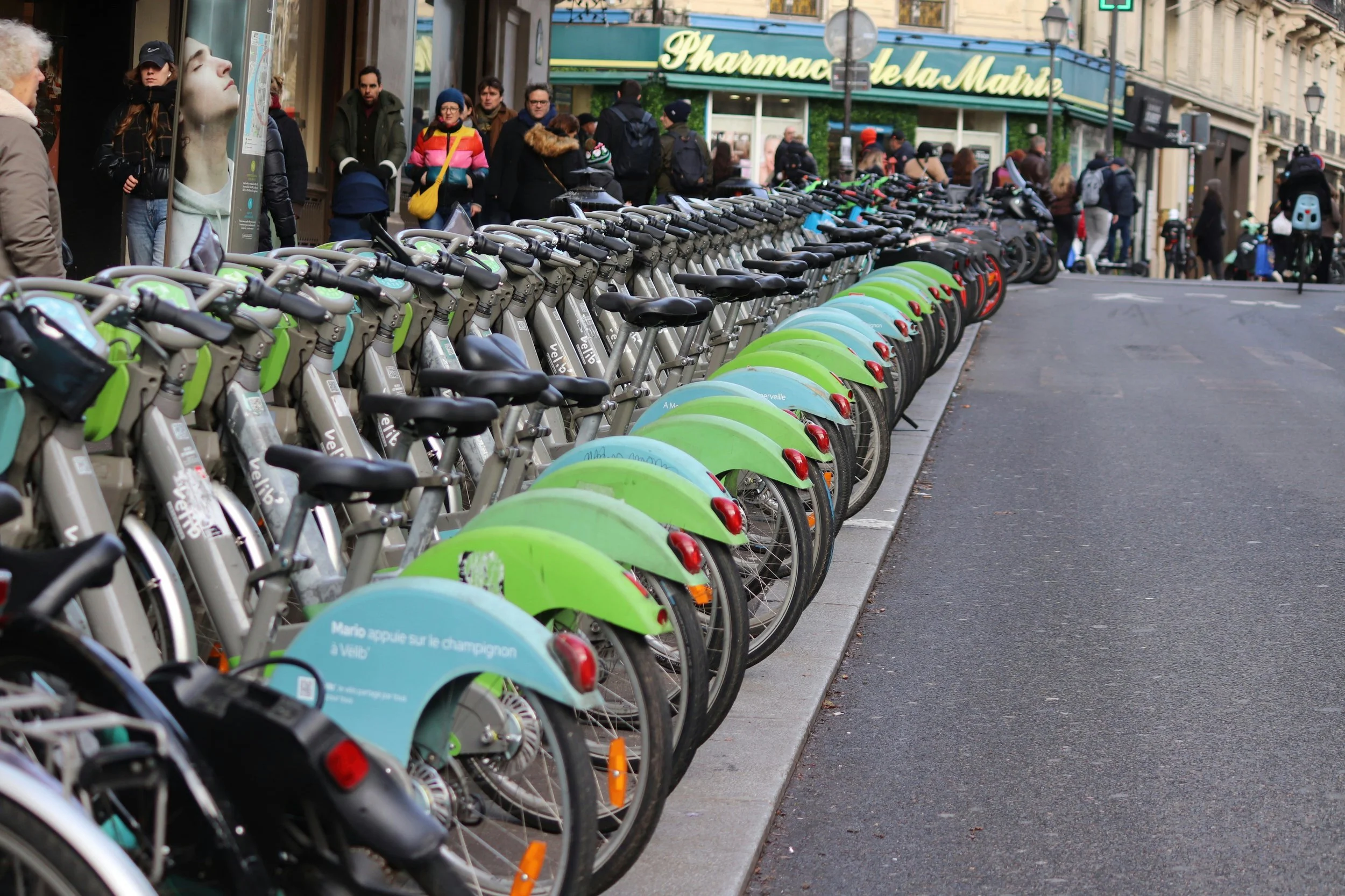 Long row of docked Vélib' bike-share bicycles along a busy Paris sidewalk near a pharmacy