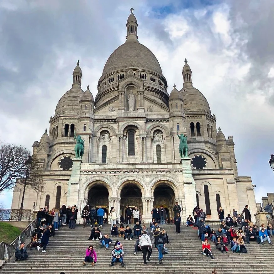 The Basilique du Sacré-Cœur de Montmartre rises above the crowd on a cloudy day, its pale Romanesque-Byzantine domes and bell-shaped turrets set against a gray-blue sky. Two green equestrian statues flank the triple-arched entrance porch. Dozens of v
