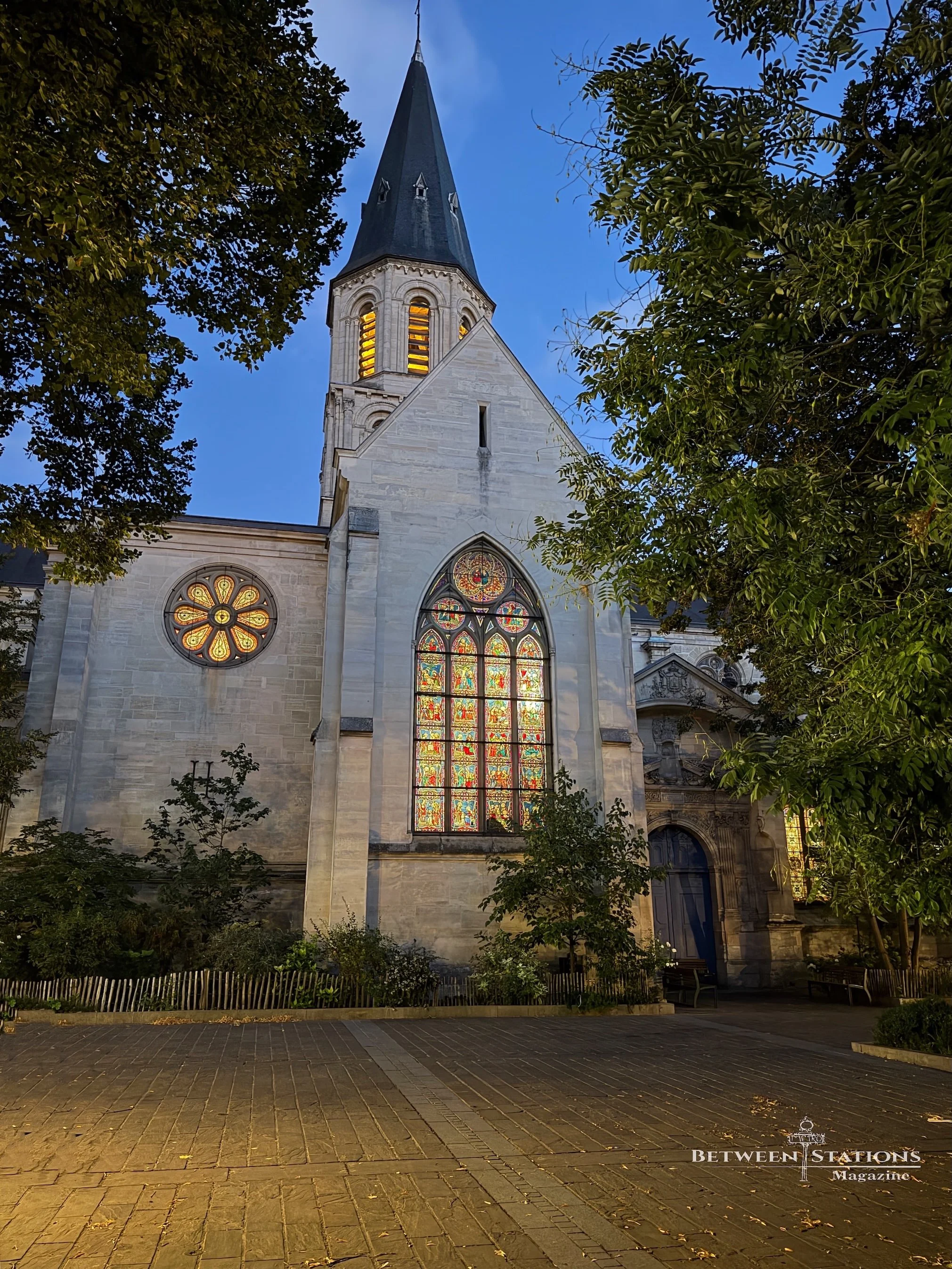 At blue hour, the Église Saint-Pierre-Saint-Paul stands behind a paved square, its slate-roofed bell tower rising into a deep blue sky. A large pointed-arch window and a round rose window glow orange, red and gold from the light inside, and the belfr