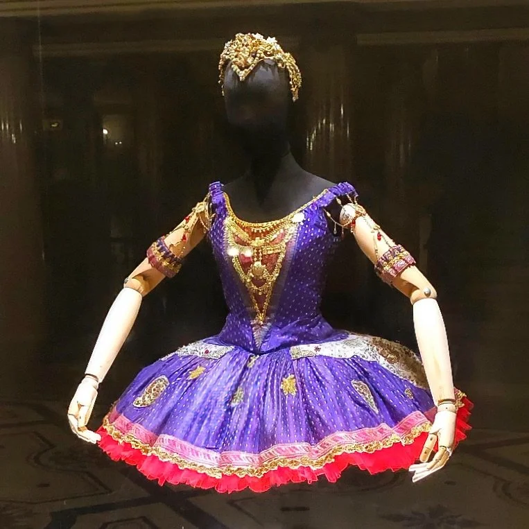 Behind glass, a mannequin with wooden articulated arms wears a ballerina costume: a purple bodice and short tutu dotted with small motifs, gold chain and jewel trim across the chest, and a pink and gold border above a ruffle of bright red tulle. Bead