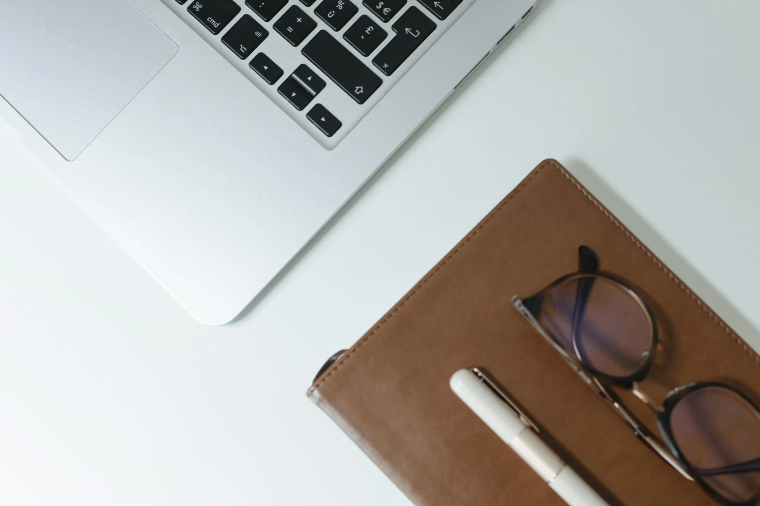Close-up of a silver laptop and a brown leather journal with glasses inside. A white pen is also visible on the journal, all placed on a white surface.
