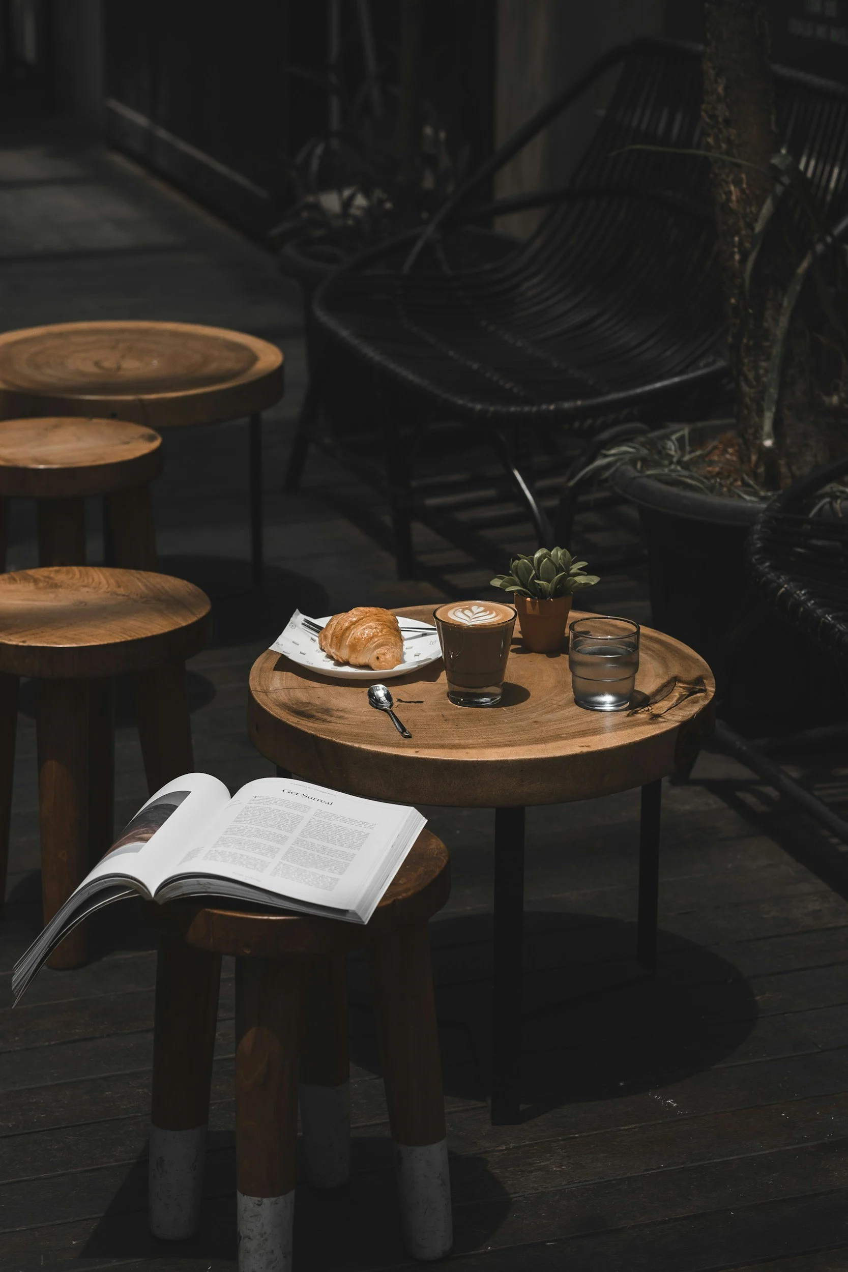 Latte, croissant, and cup of water on a circular wooden table, with an open book on a nearby seat.