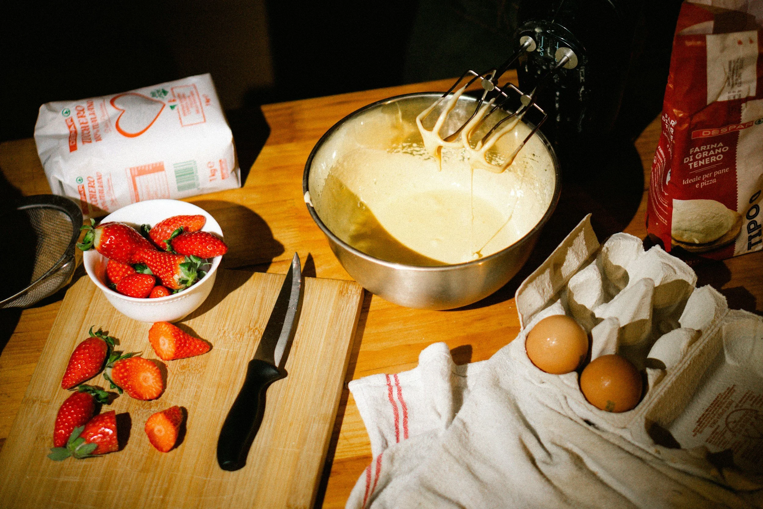 Baking implements with strawberries, cutting board with knife, mixing bowl with batter, carton of eggs.