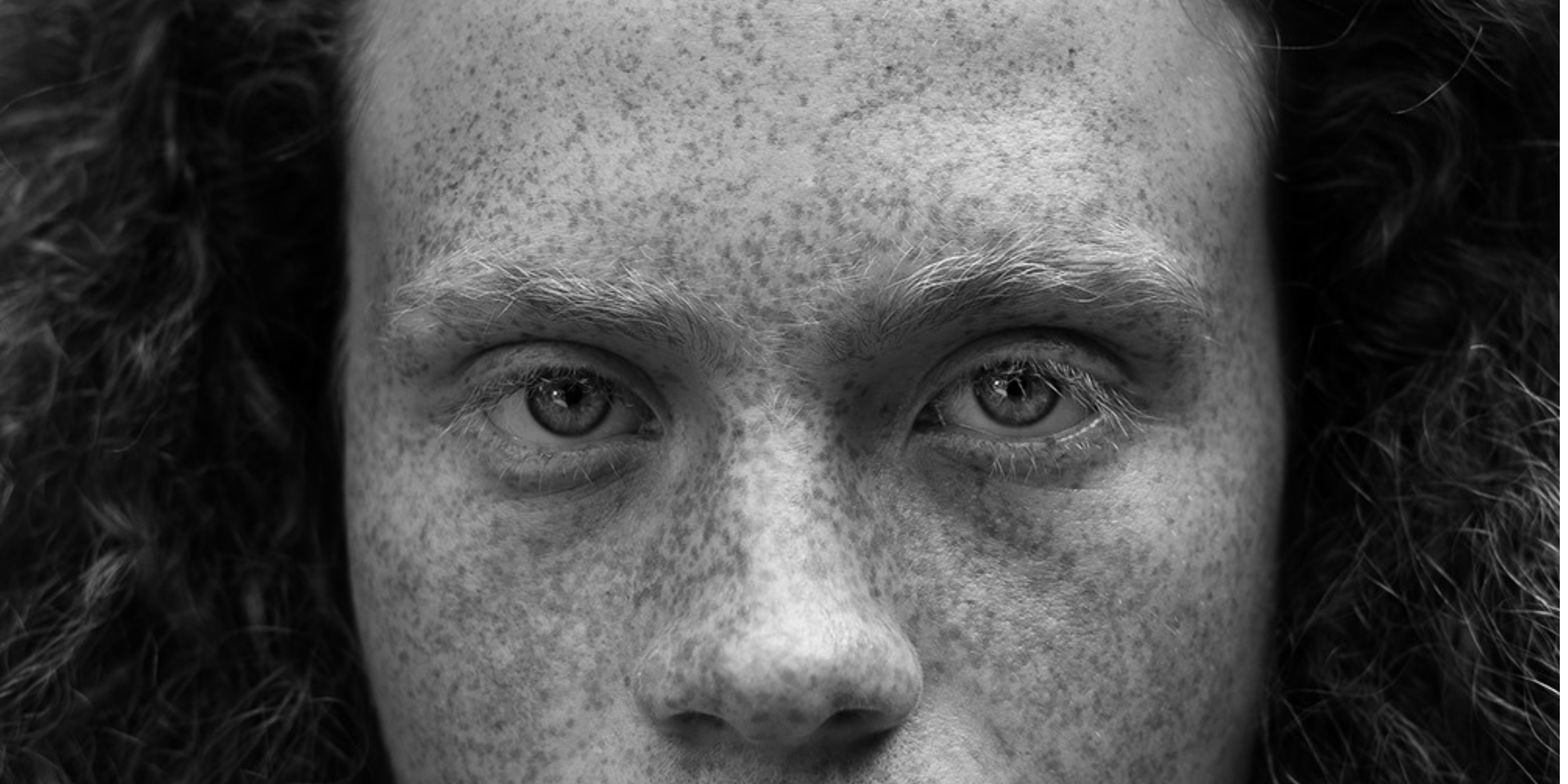 Close-up of a person's face with curly hair and freckles, focusing on their piercing eyes.