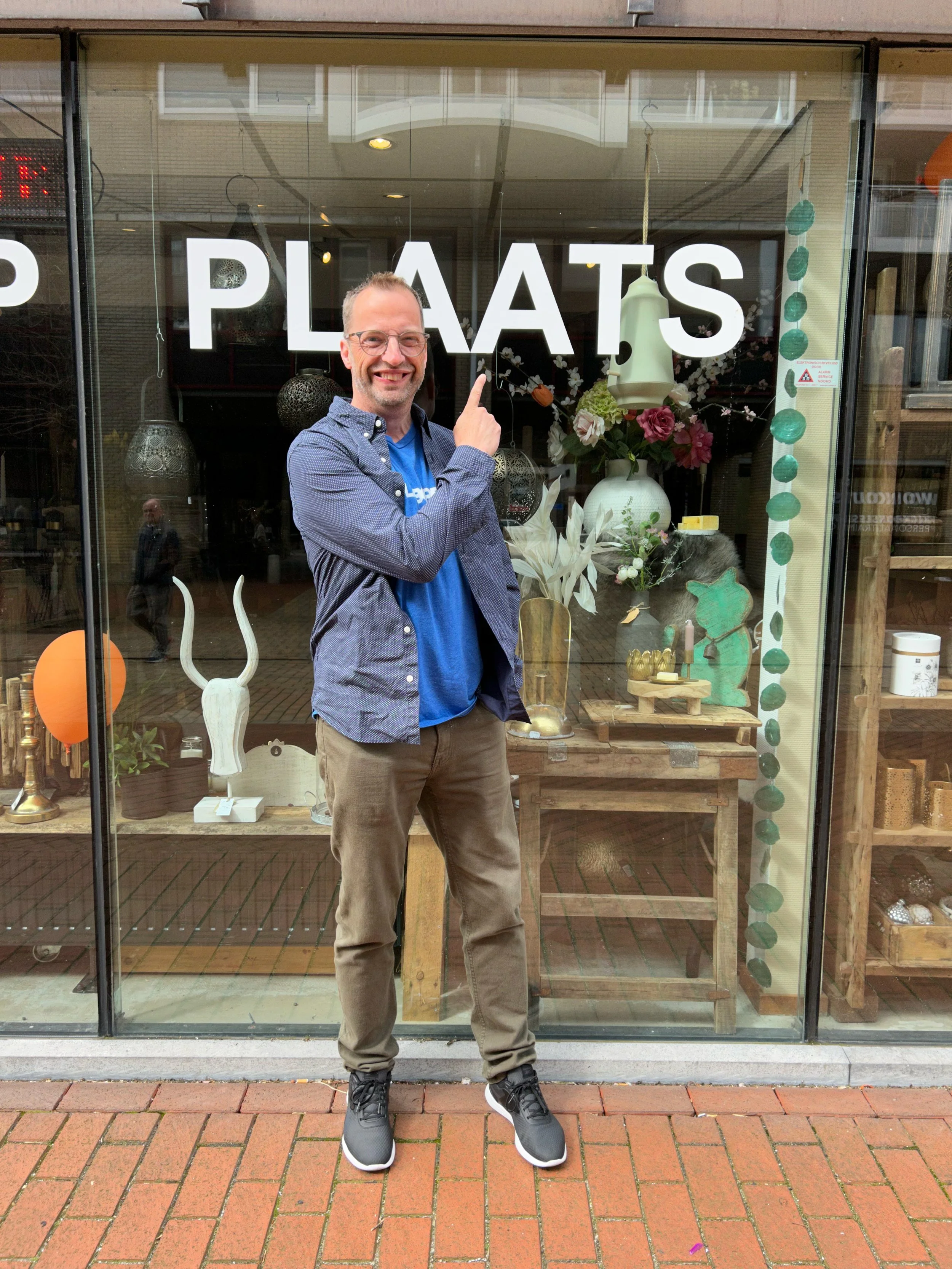 Smiling man standing outside a store window with decorative items and the word 'PLATS' written on the glass, pointing at the text.