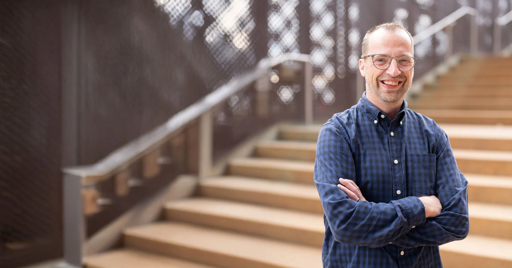 A man with glasses, wearing a blue plaid shirt, smiling with arms crossed, standing in front of a staircase with wooden steps and metal railing.