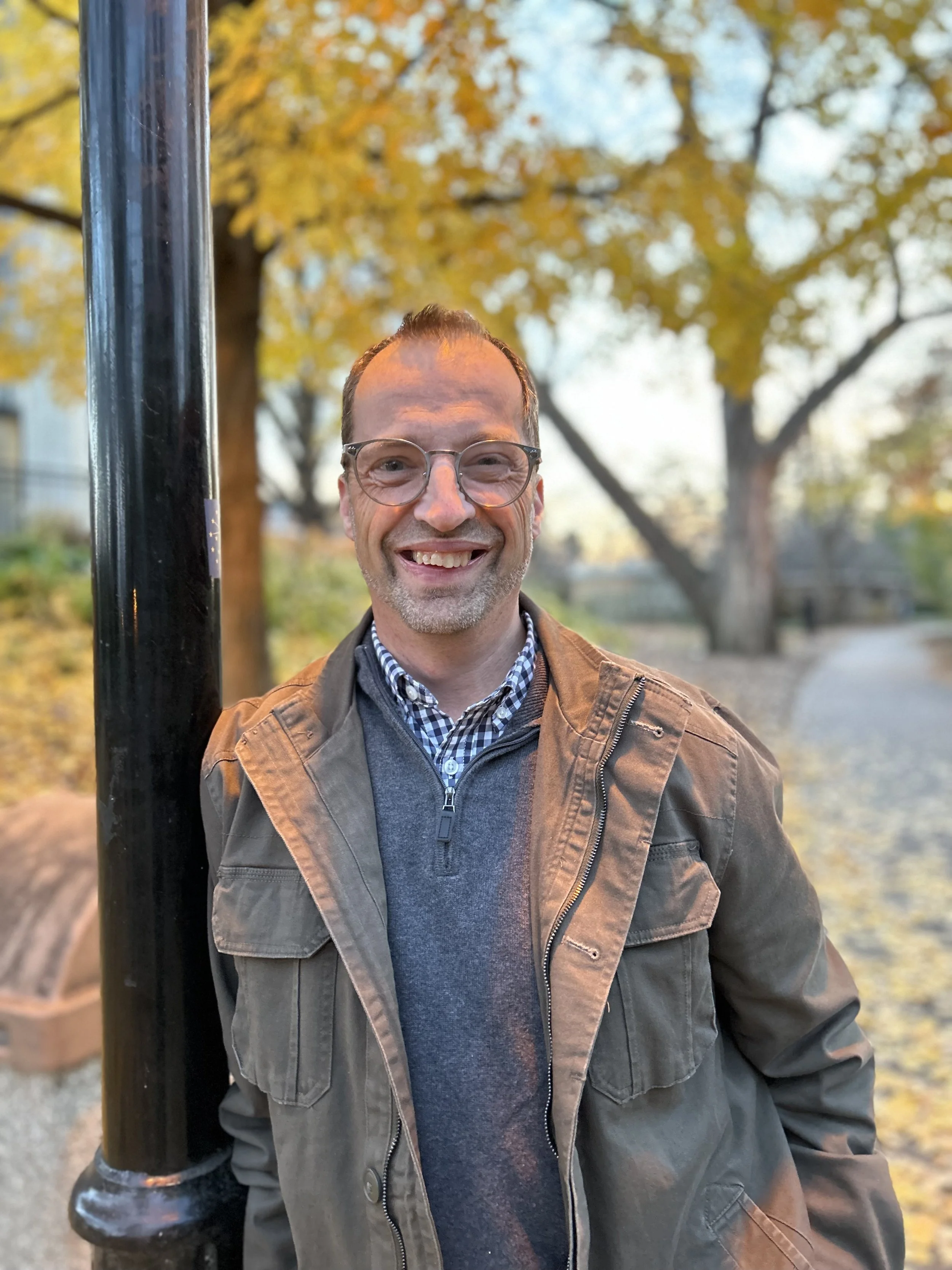 A man smiling outdoors during fall with yellow and orange leaves on trees behind him, wearing glasses, a checkered shirt, gray sweater, and a brown jacket, standing next to a black pole.