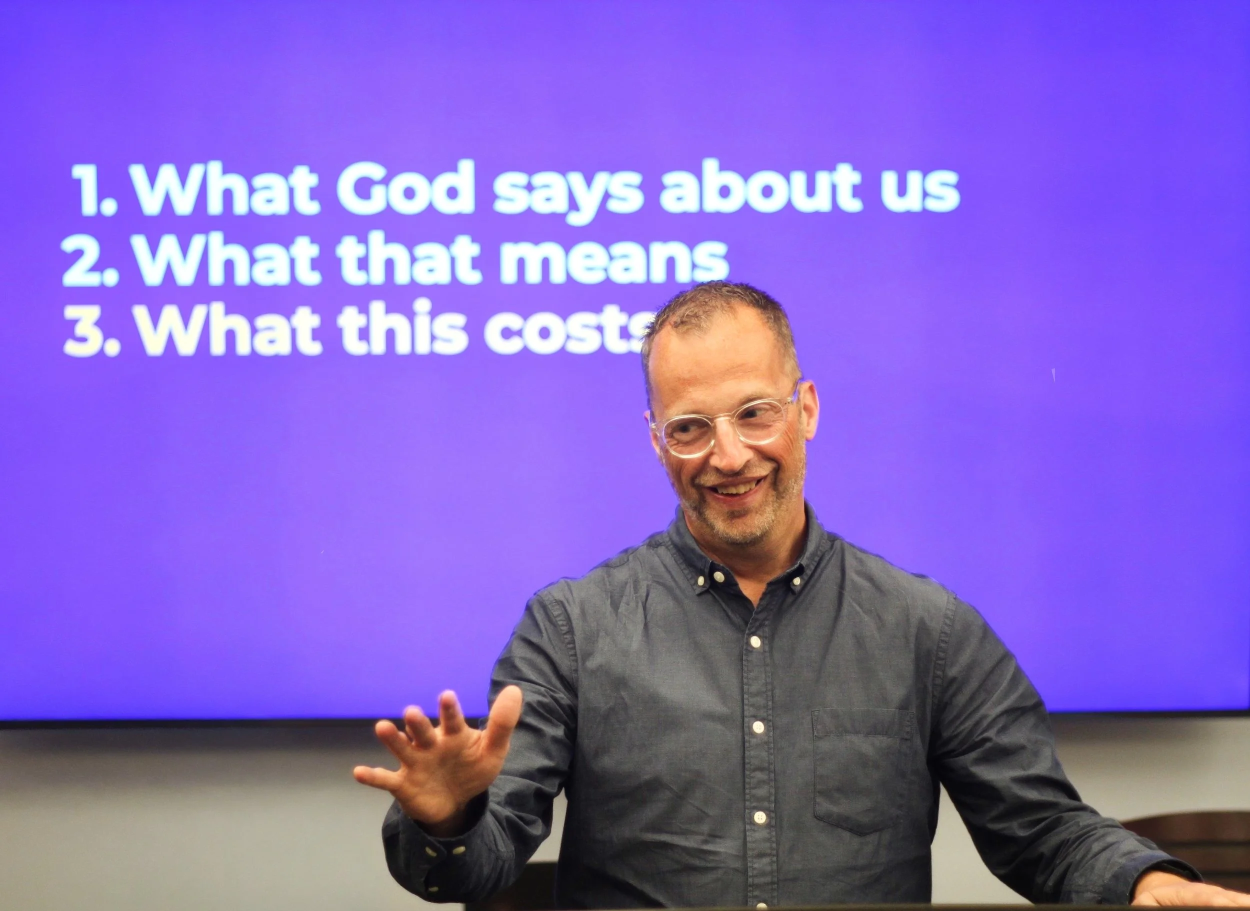 A man with glasses and a gray button-up shirt smiling and gesturing with his hand in front of a purple screen that displays a numbered list.