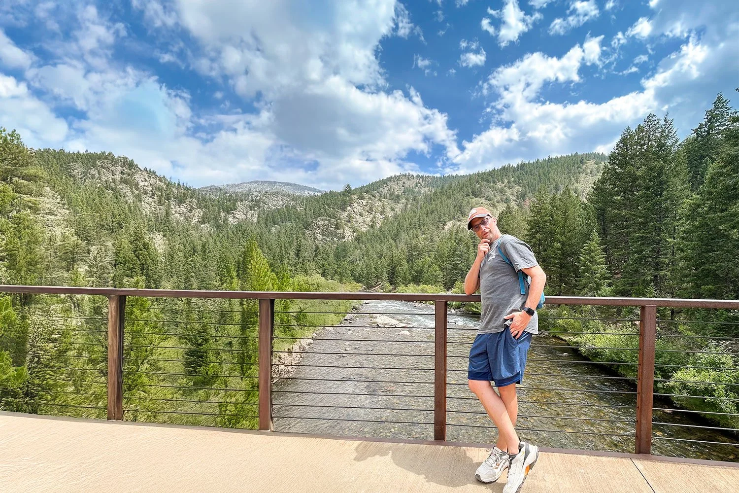 Man standing on a bridge overlooking a forested canyon with a river below, mountains in the background, and a partly cloudy sky overhead.