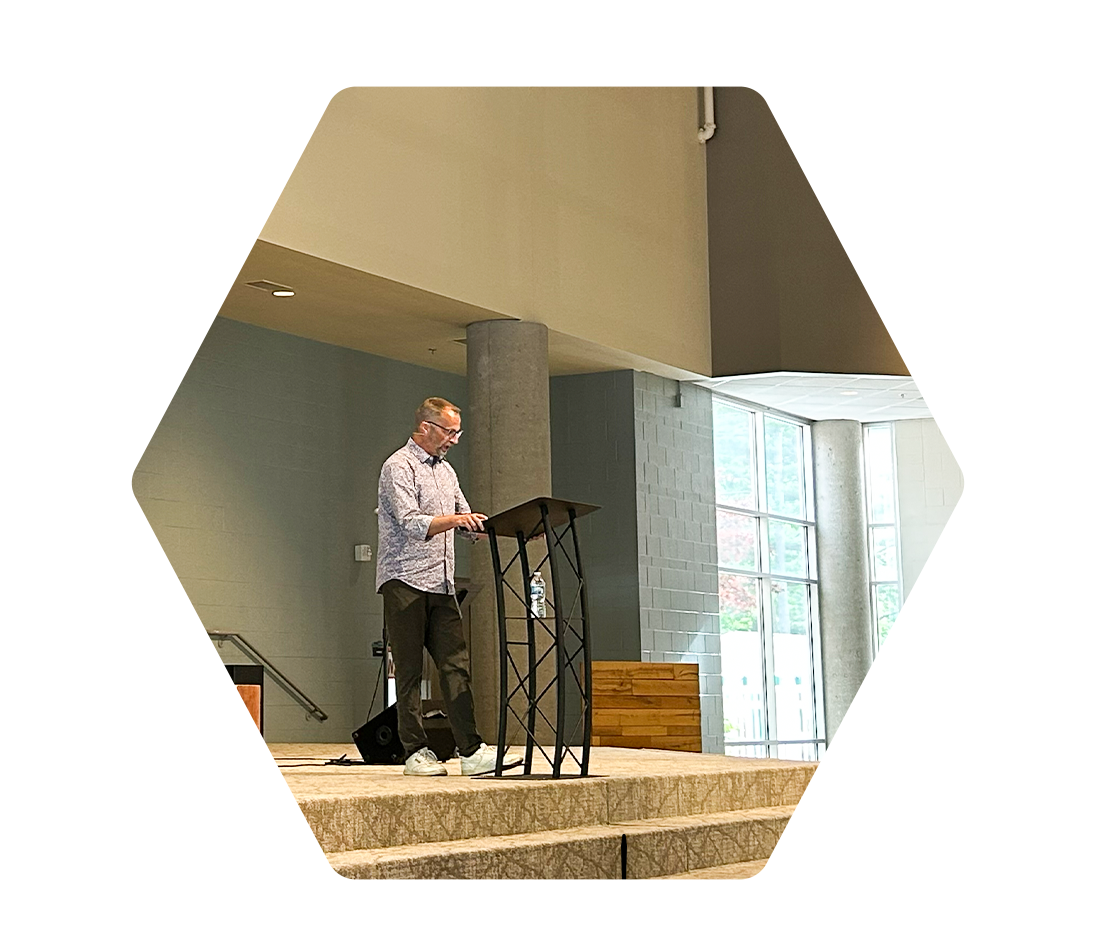 A man at a podium giving a speech or presentation in a large, modern indoor space with floor-to-ceiling windows.