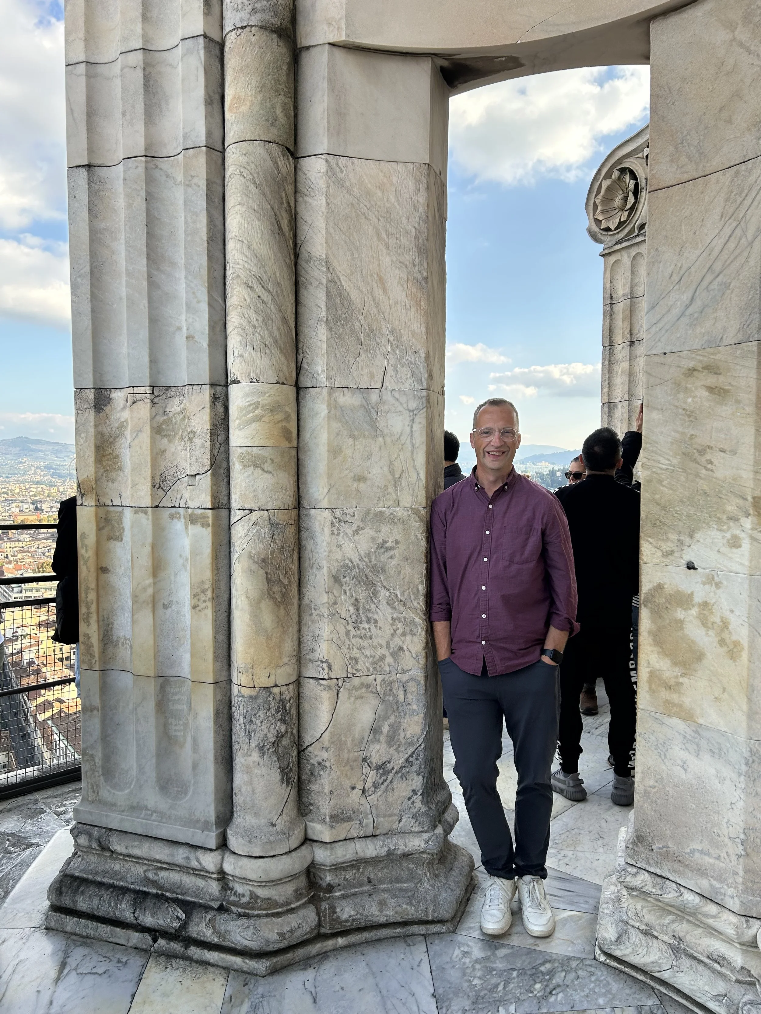 A man in a purple shirt and dark pants standing on marble floor inside a historic marble structure with large marble columns featuring cracks, overlooking a cityscape with mountains in the distance under a partly cloudy sky.