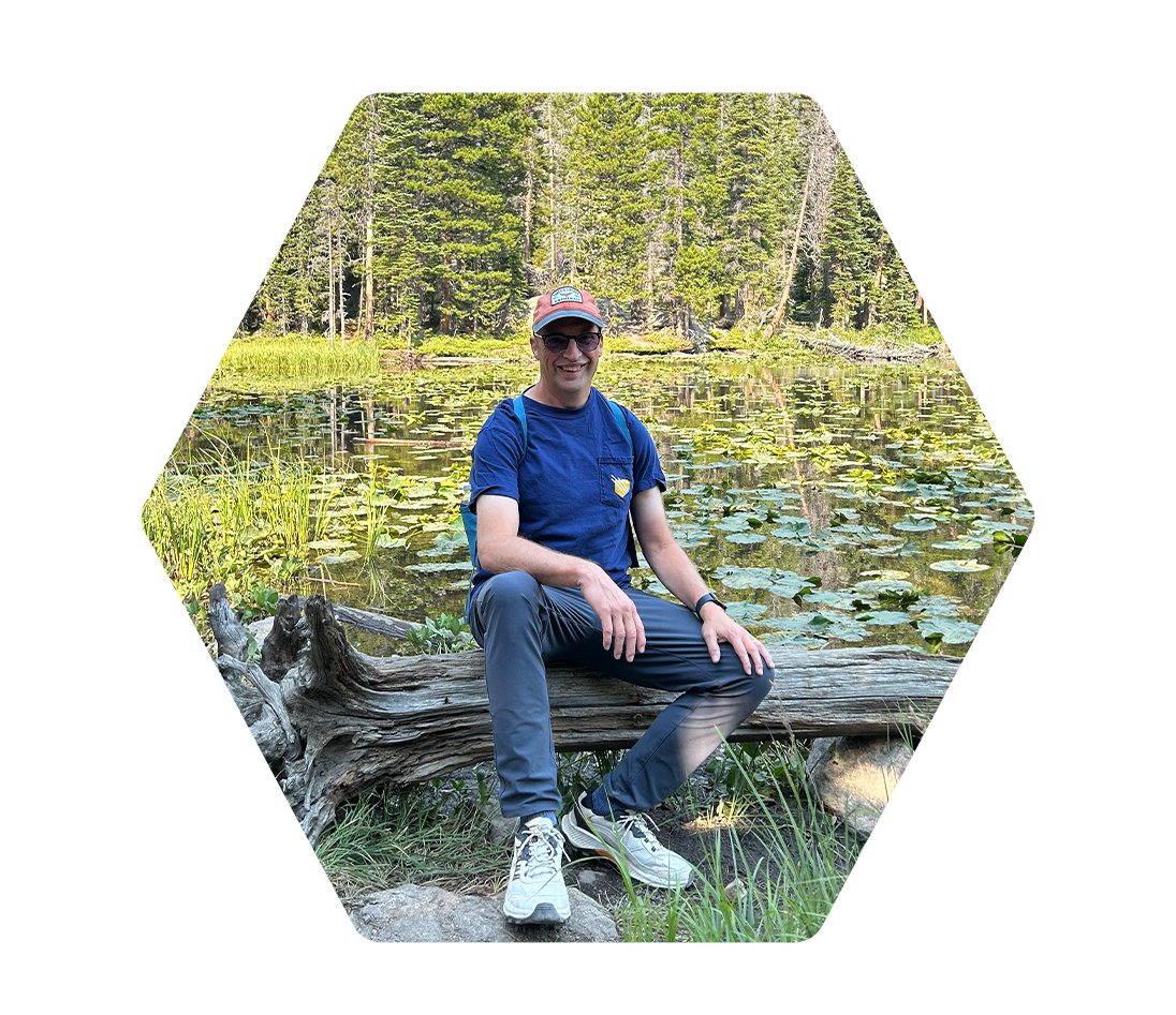 A man smiling and sitting on a fallen log at a pond with lily pads, surrounded by trees in a forest.
