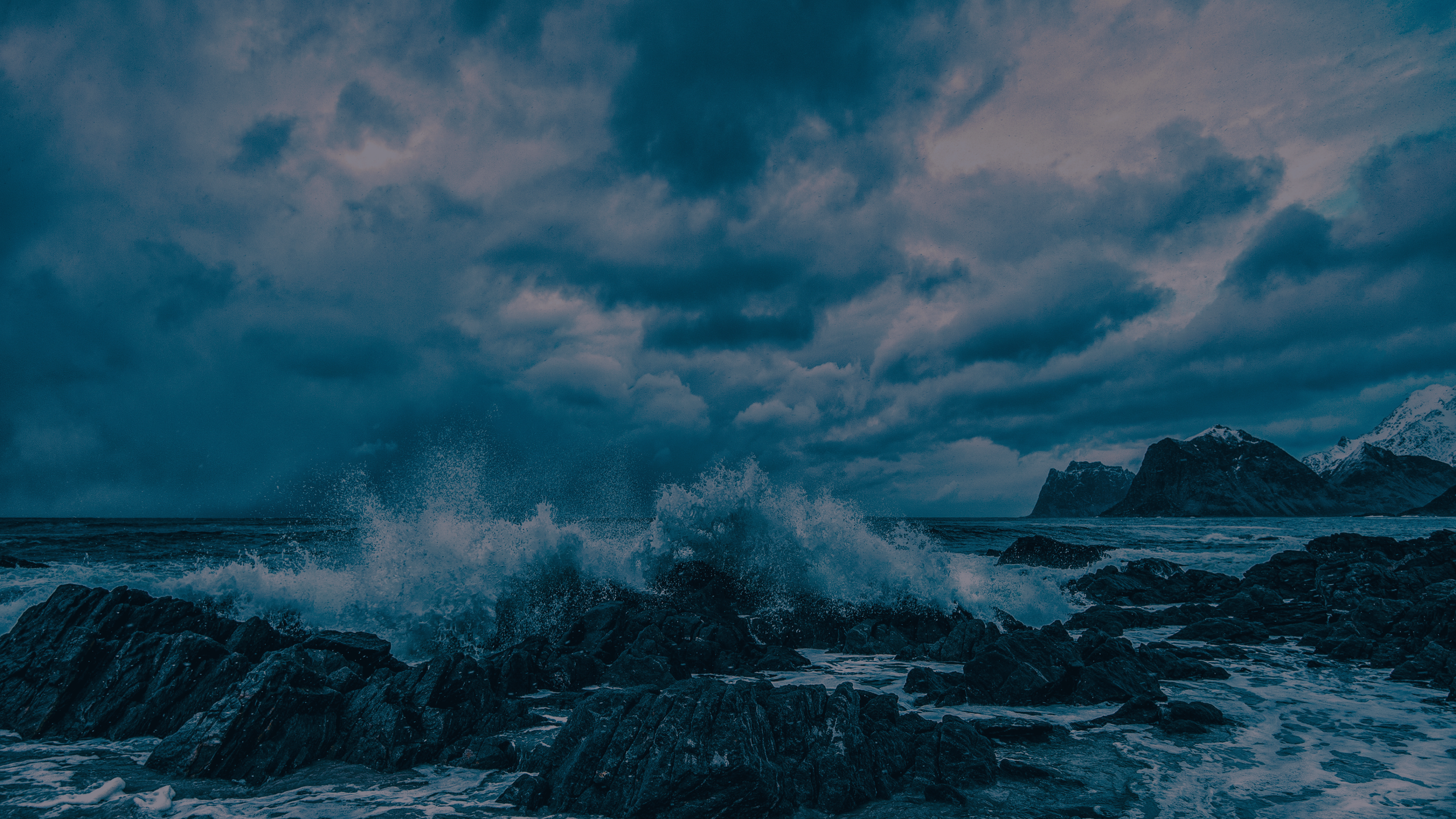 Overcast sky with dark clouds over a rocky coastline with waves crashing against rocks in the ocean.
