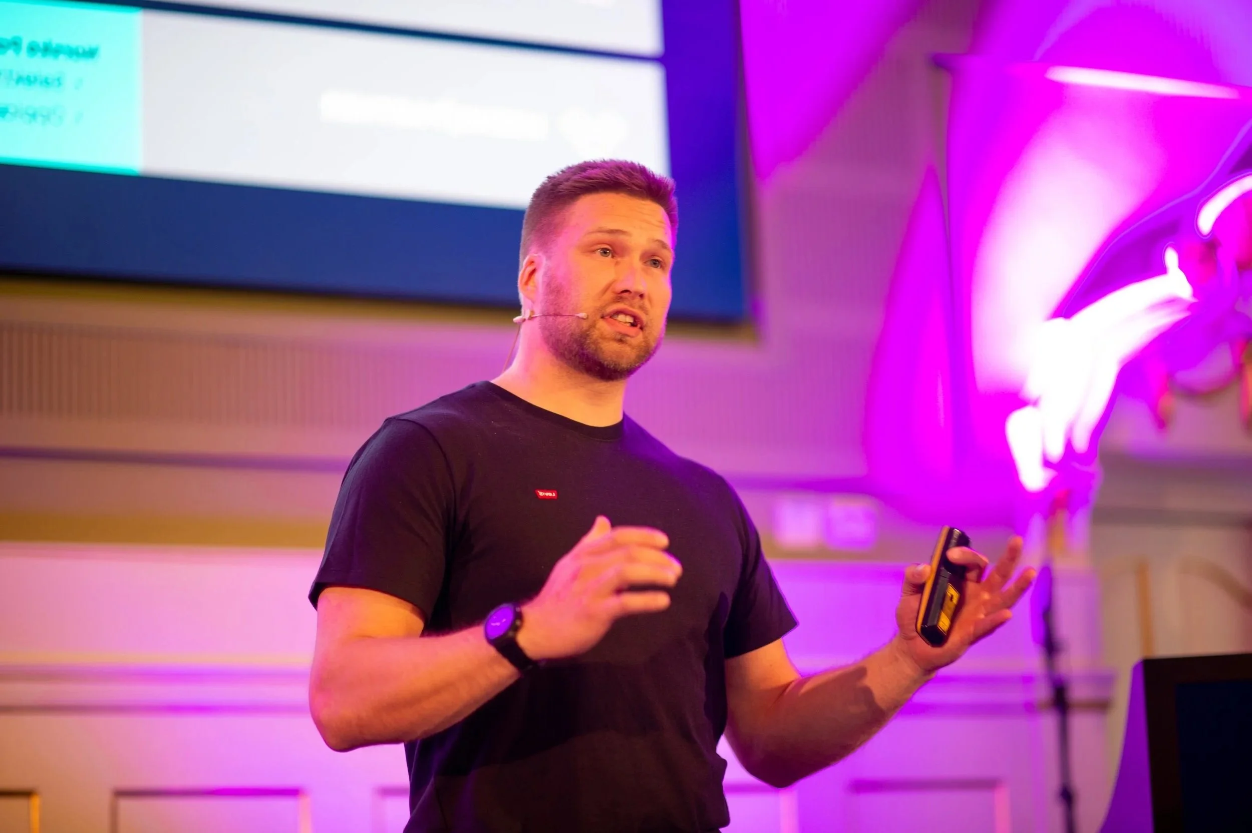 Krister Halonen giving a presentation about innoveks and Reinventing Underwater Concrete Repair in a room lit with pink and purple lights. He is wearing a black T-shirt, a watch, and holding a microphone and a small device.