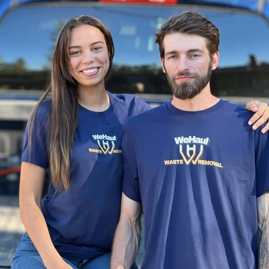 Two people standing outdoors in front of a truck, both wearing blue t-shirts with a logo and the words "WeHaul Waste Removal." The woman has long brown hair and is smiling, while the man has short brown hair, a beard, and a neutral expression. The woman has her arm around the man's shoulder.