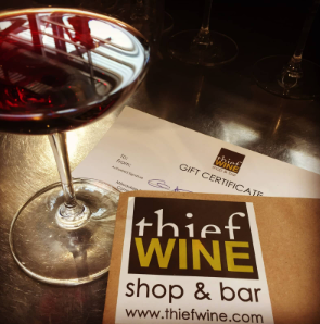 A glass of red wine next to a Thief Wine Shop and Bar in Milwaukee gift certificate on a dark wooden table.
