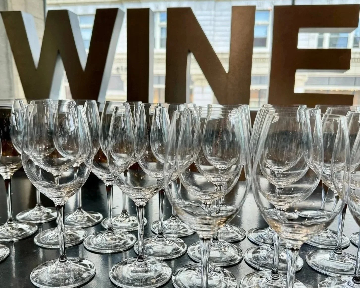 Multiple empty wine glasses arranged on a table with a large 'WINE' sign in the background at Thief Wine Shop and Bar's Grand Tasting in the Milwaukee Public Market.
