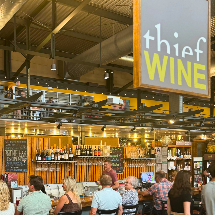 People sitting at Thief Wine Shop and Bar with shelves of wine bottles behind the bar and a sign overhead that reads 'Thief Wine'  in Milwaukee Public Market in the Historic Third Ward of Milwaukee, Wisconsin