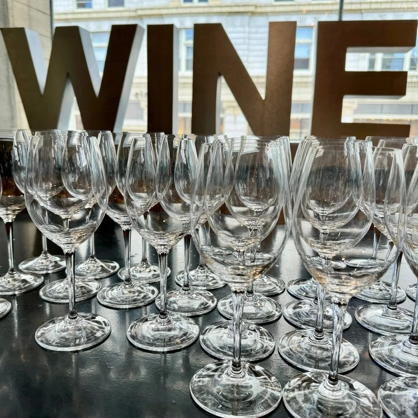 Multiple empty wine glasses arranged on a table with a large 'WINE' sign in the background at Thief Wine Shop and Bar's Grand Tasting in the Milwaukee Public Market in the Historic Third Ward.