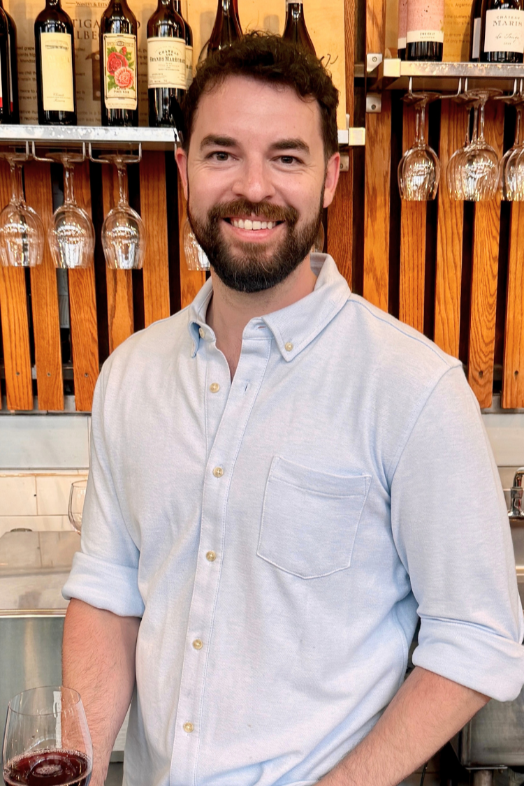 Jordan Phillips, manager at Thief Wine Shop and Bar, standing in front of a bar with wine bottles and hanging wine glasses.