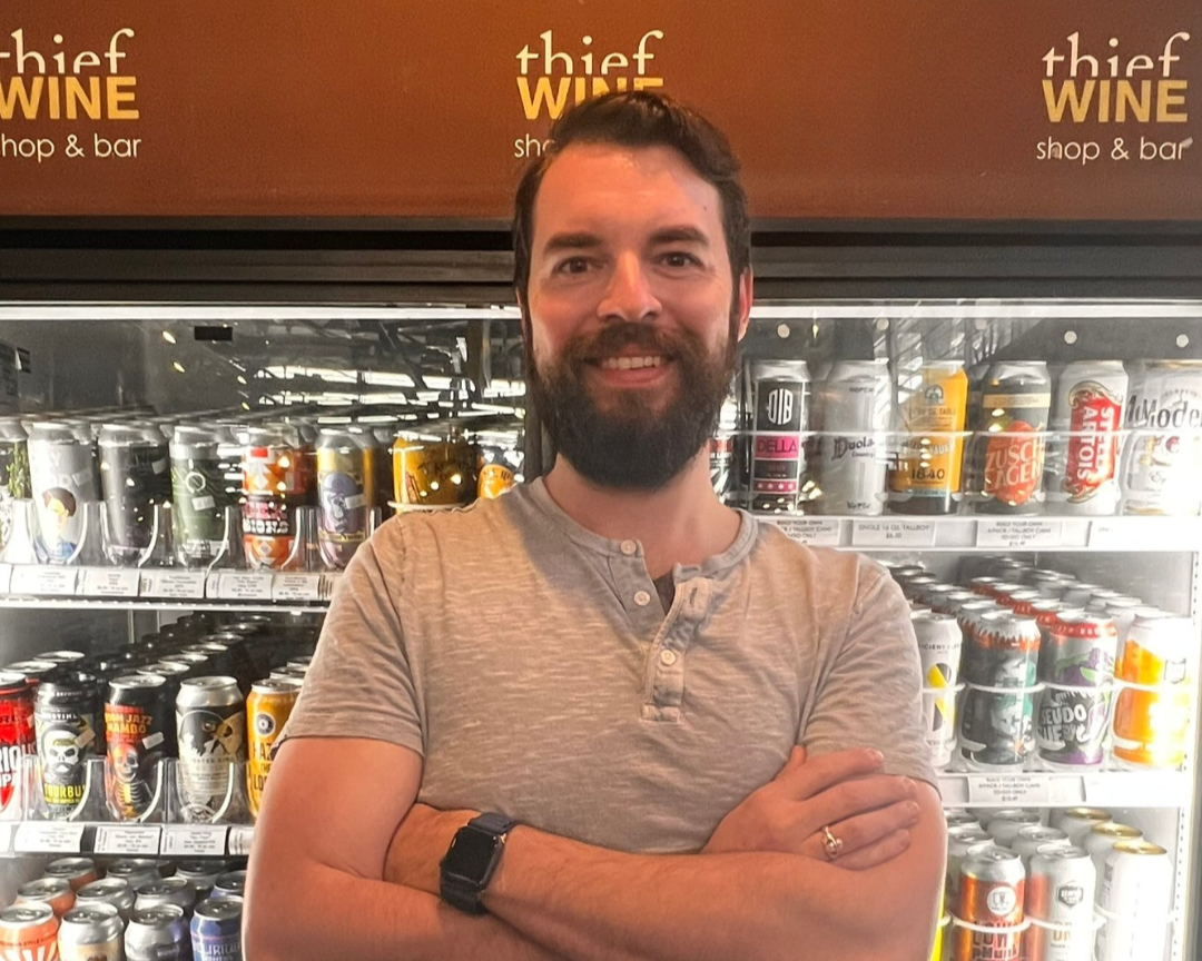 Jordan Phillips, Thief Wine Shop and Bar manager smiling with arms crossed, standing in front of a fridge stocked with beer cans.