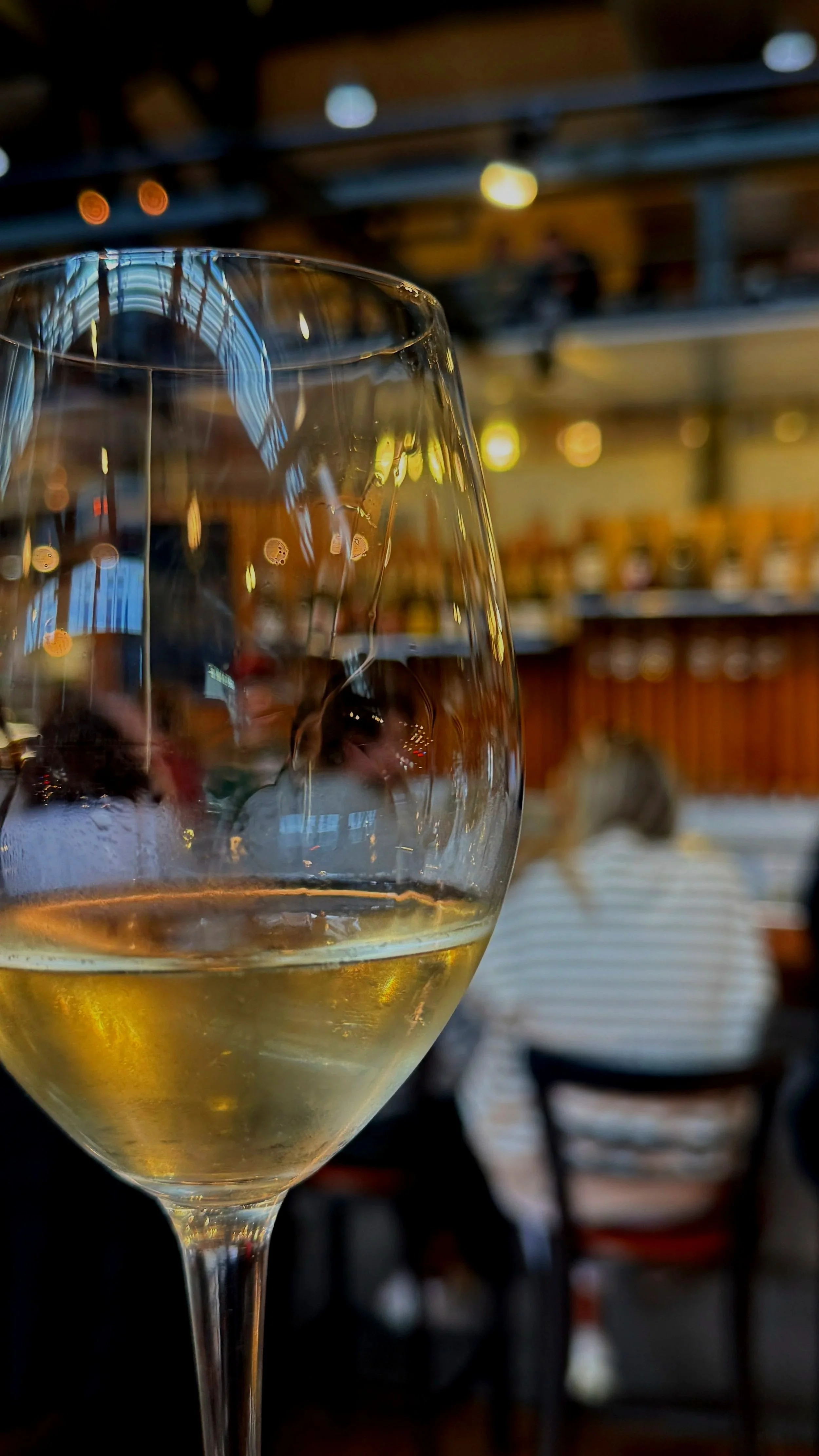 A glass of white wine in the foreground with a blurry background of Thief Wine Shop and Bar, including people sitting and a wooden bar counter.
