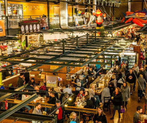 Crowded indoor shot of the Milwaukee Public Market with various food stalls, shoppers, and overhead lighting.
