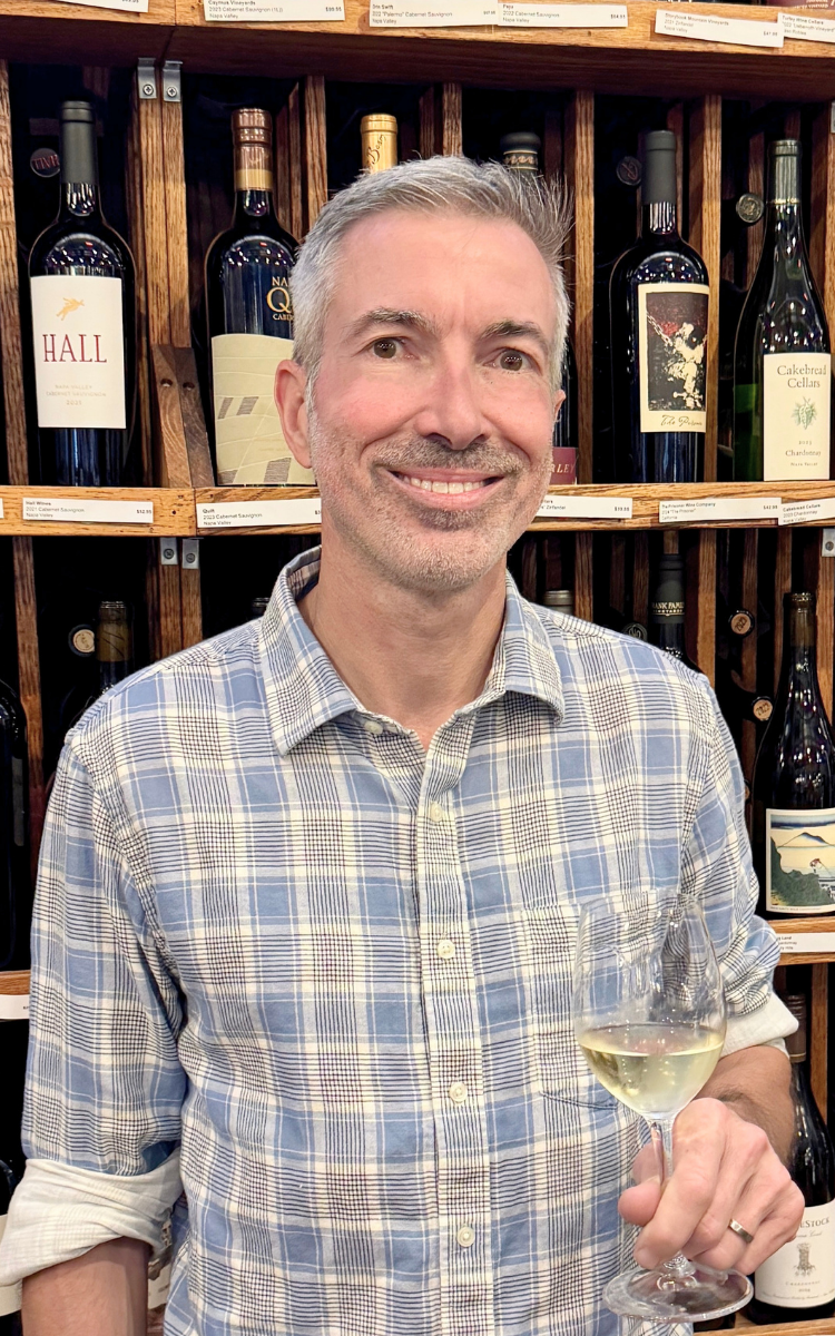 Phil Bilodeau, owner of Thief Wine Shop and Bar holding a glass of white wine, standing in front of a shelf filled with various bottles of wine.