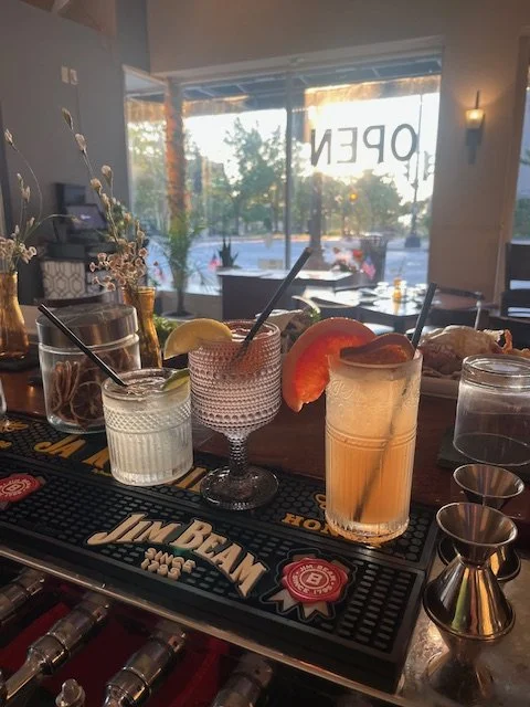 Four cocktails, mocktails, garnishes, such as  lemon, lime, and orange slices garnished on a bar counter in front of a window. Fresh fruit, good drinks and fun times. Happy hour.