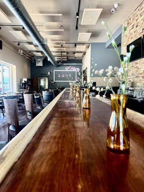 A bar or restaurant interior with a long wooden counter decorated with small vases of white flowers, and a seating area with chairs and tables in the background. Exposed ductwork and modern ceiling design are visible.