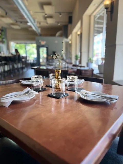 A restaurant table set with white plates, napkins, glasses of water, and a centerpiece with flowers in a gold vase, inside a well-lit, modern dining area.