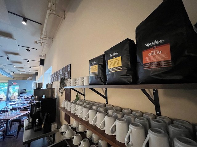 Shelves with bags of coffee labeled "Brazil Decaf" and rows of white mugs in a cafe or coffee shop.