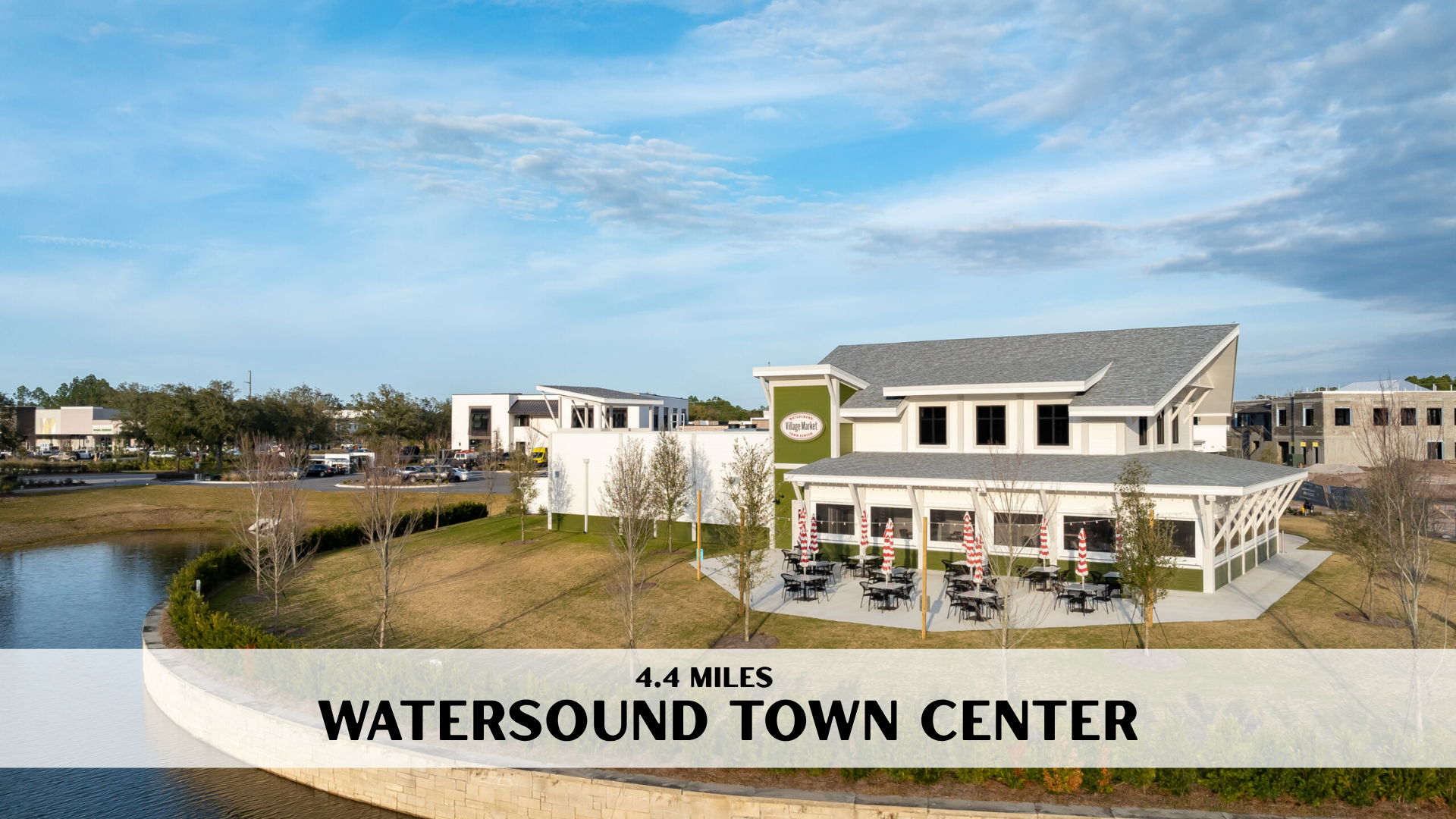 A modern shopping center called Watersound Town Center with outdoor seating and umbrellas, near a pond and landscaped grassy area, under a partly cloudy sky.