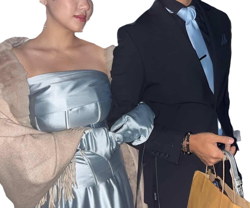 Close-up of a woman and a man dressed in formal attire, holding hands. The woman wears a shiny silver strapless dress with matching gloves and a beige shawl. The man wears a black suit with a light blue shirt and bow tie, holding shopping bags.