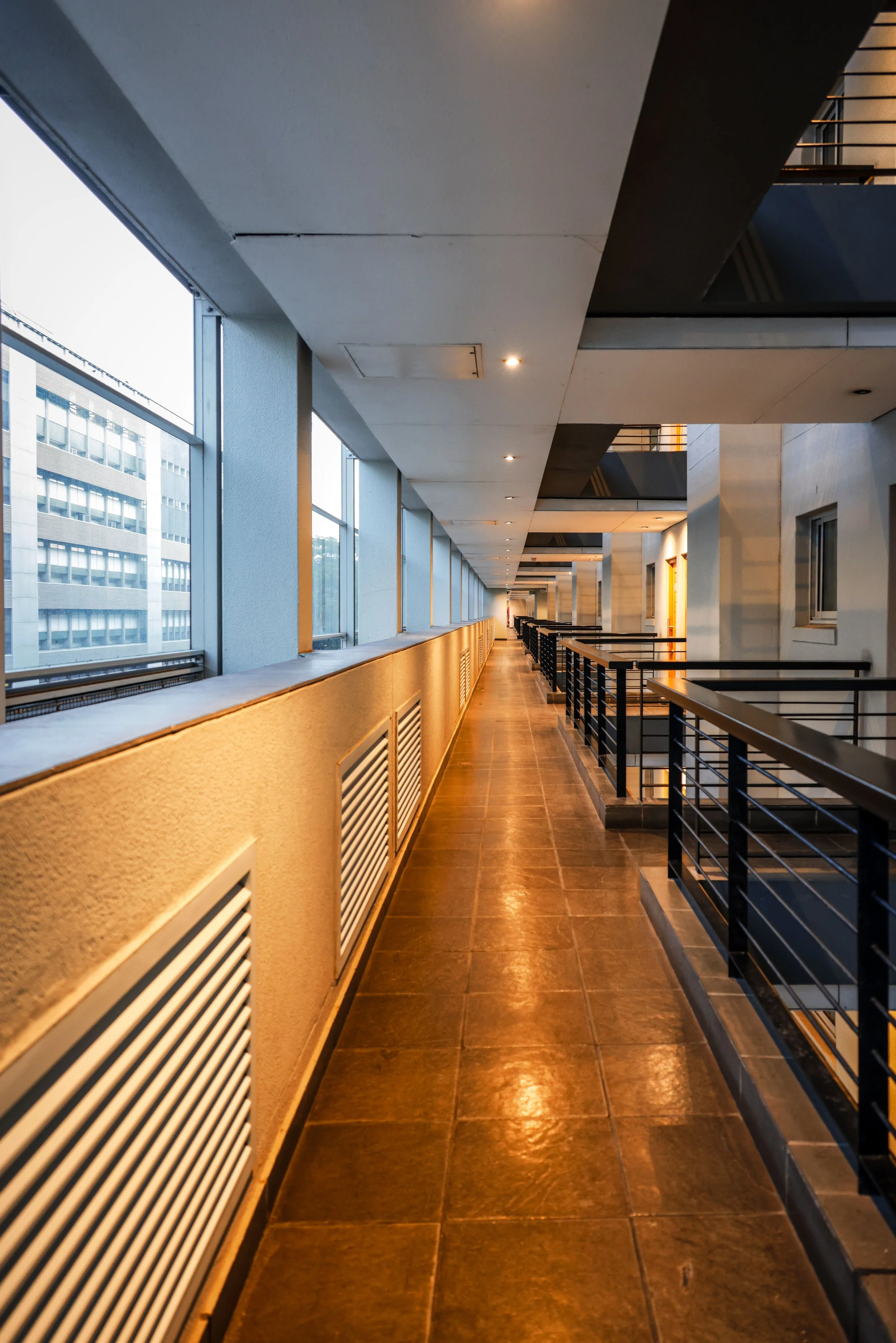 A hallway in a modern building with large windows on the left letting in natural light, beige walls, and brown tiled flooring. The hallway features black railings and multiple small ceiling lights.