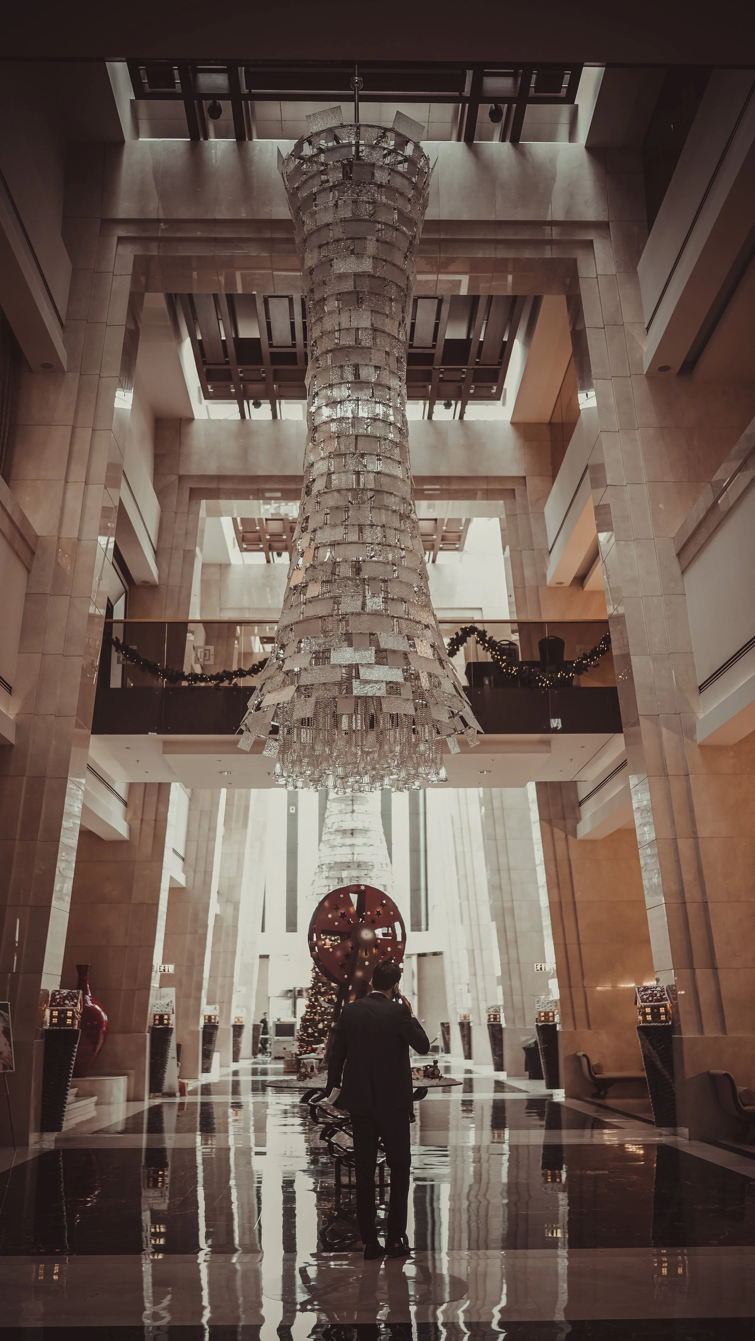 A spacious hotel lobby with a large, elaborate crystal chandelier hanging from the ceiling. The area features marble floors, decorative walls, and a Christmas tree in the background, with holiday garlands and ornaments. A man in a suit is standing near a bench, talking on his phone.