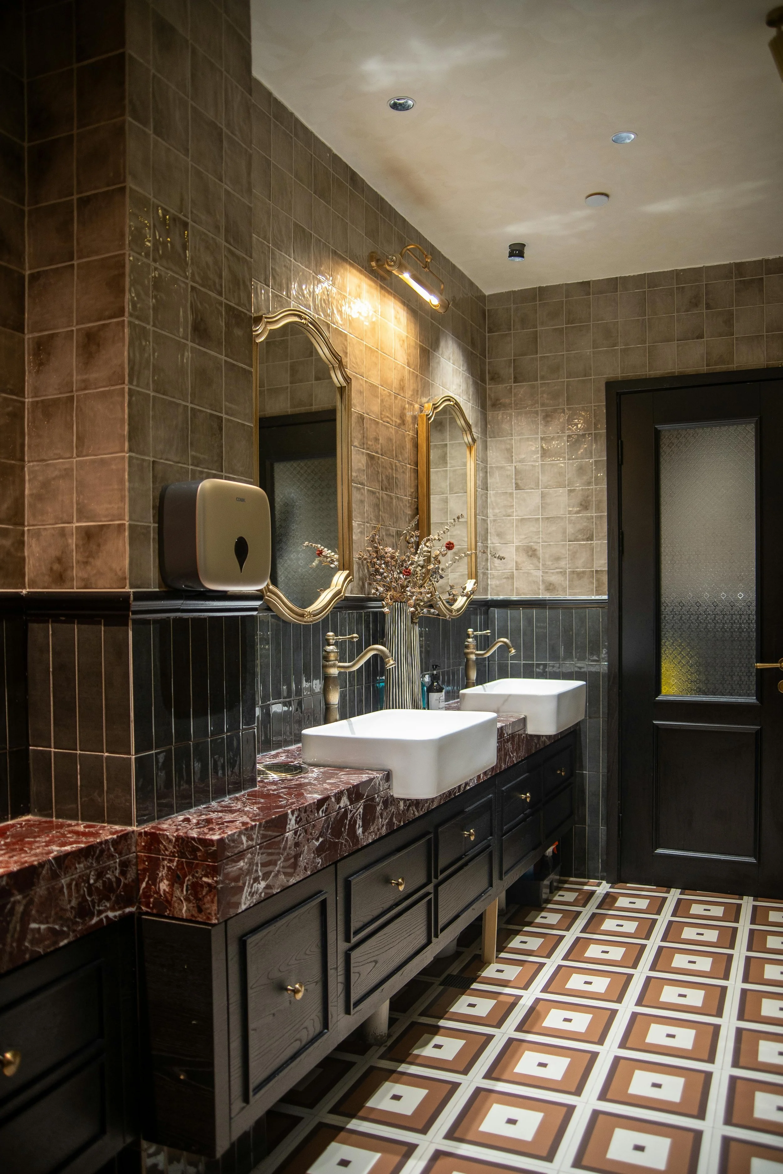 Bathroom with brown tiled walls, gold framed mirrors, a red marble countertop with two white sinks, black cabinetry, patterned tile flooring, and a black door with textured glass