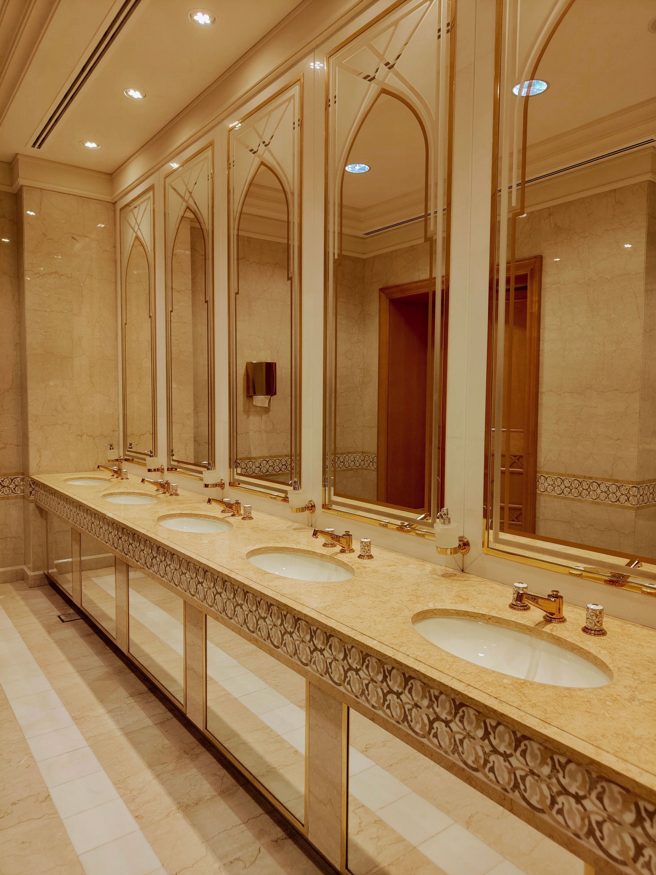 A bathroom with a long marble counter featuring six sinks and six gold-colored faucets. Large ornate mirrors are mounted above each sink, and the wall behind is decorated with beige marble tiles and decorative trim. There are ceiling lights and a tissue dispenser on the wall.