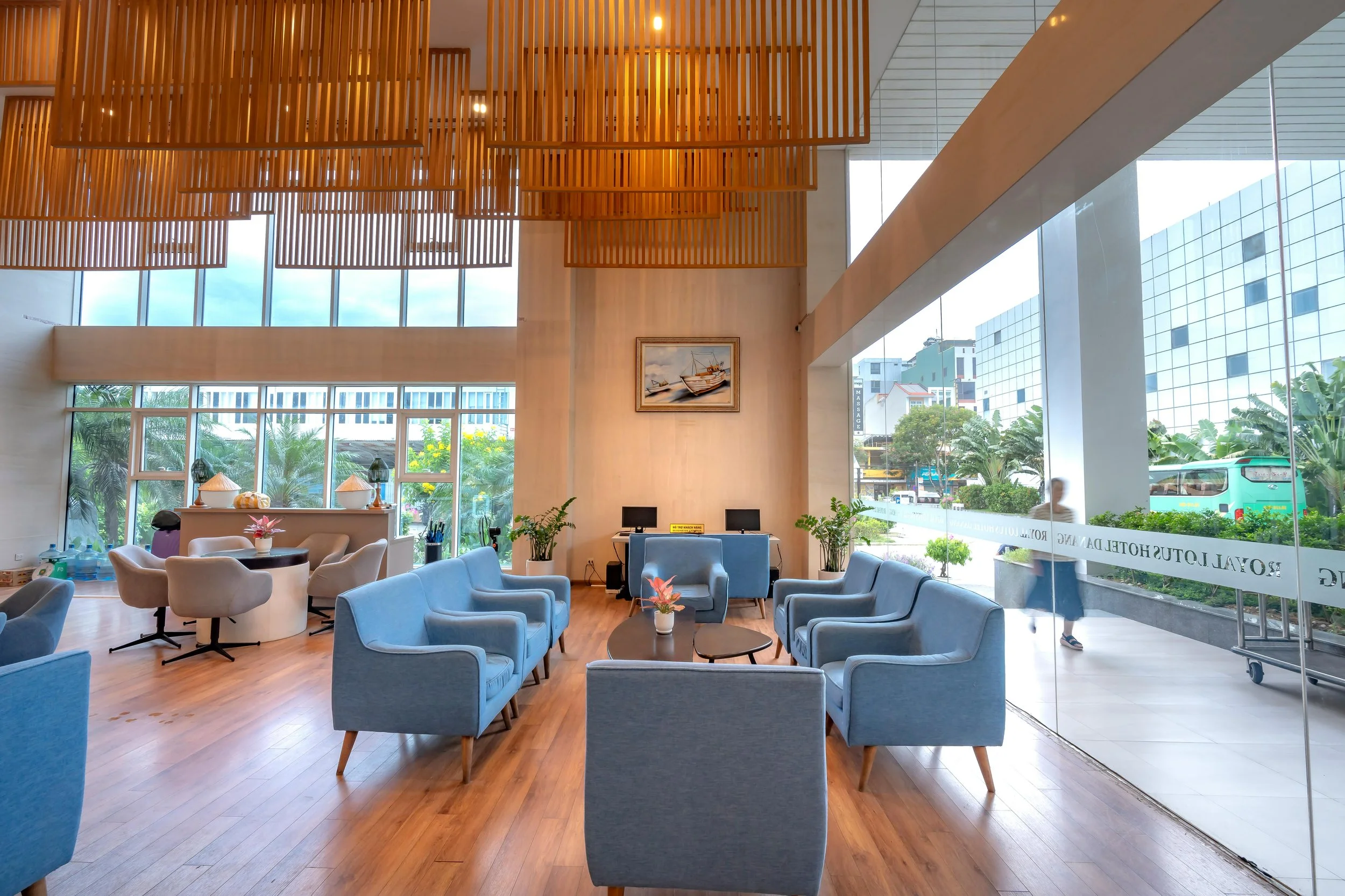 Interior of a modern lobby with blue armchairs, small round tables with flower vases, large windows, and wooden ceiling decor.